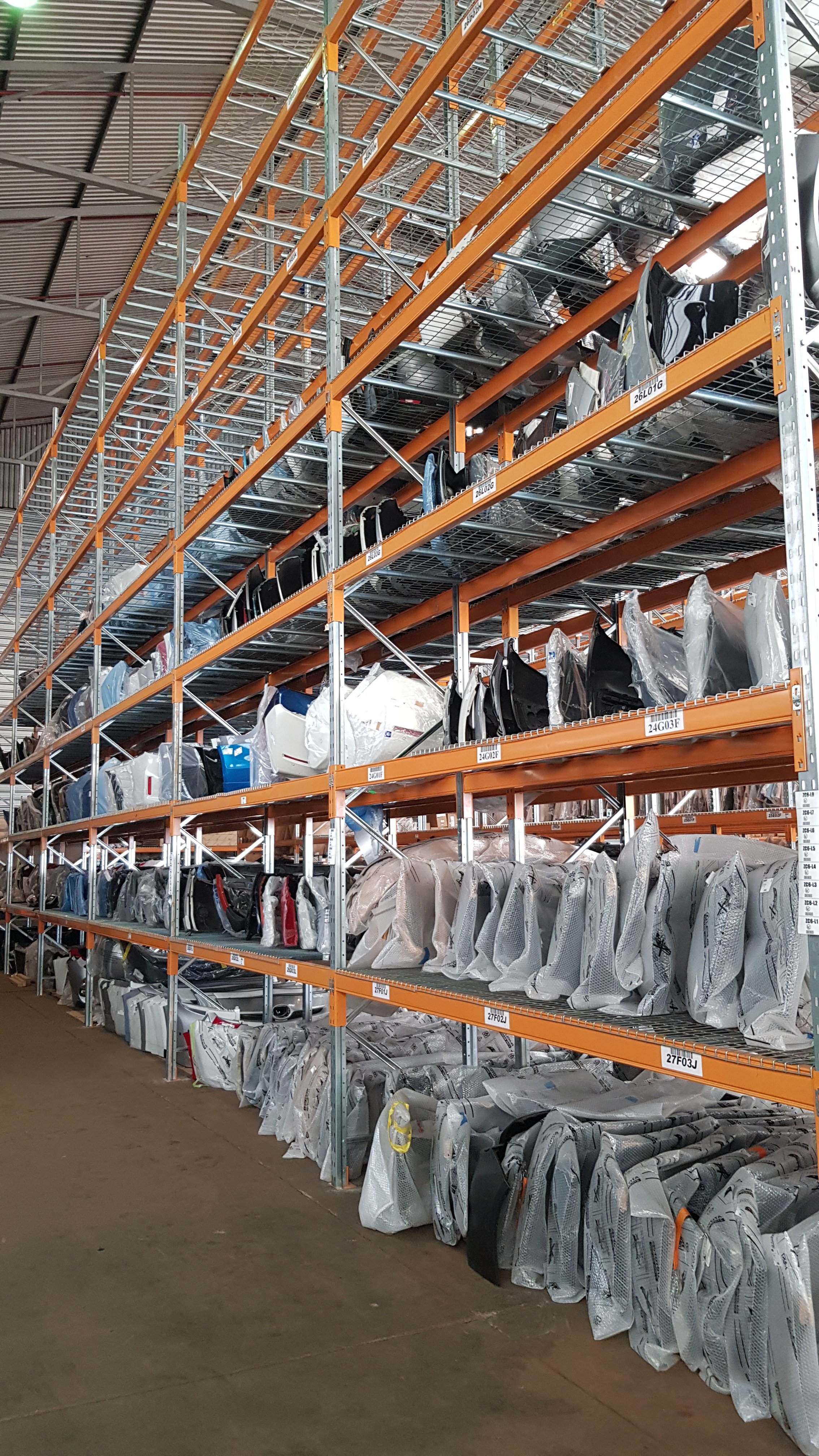 View of a large warehouse storage area with metal shelves stocked with automotive parts, primarily car doors, wrapped in plastic. The shelves are organized with labels and the aisle is visible in the foreground.