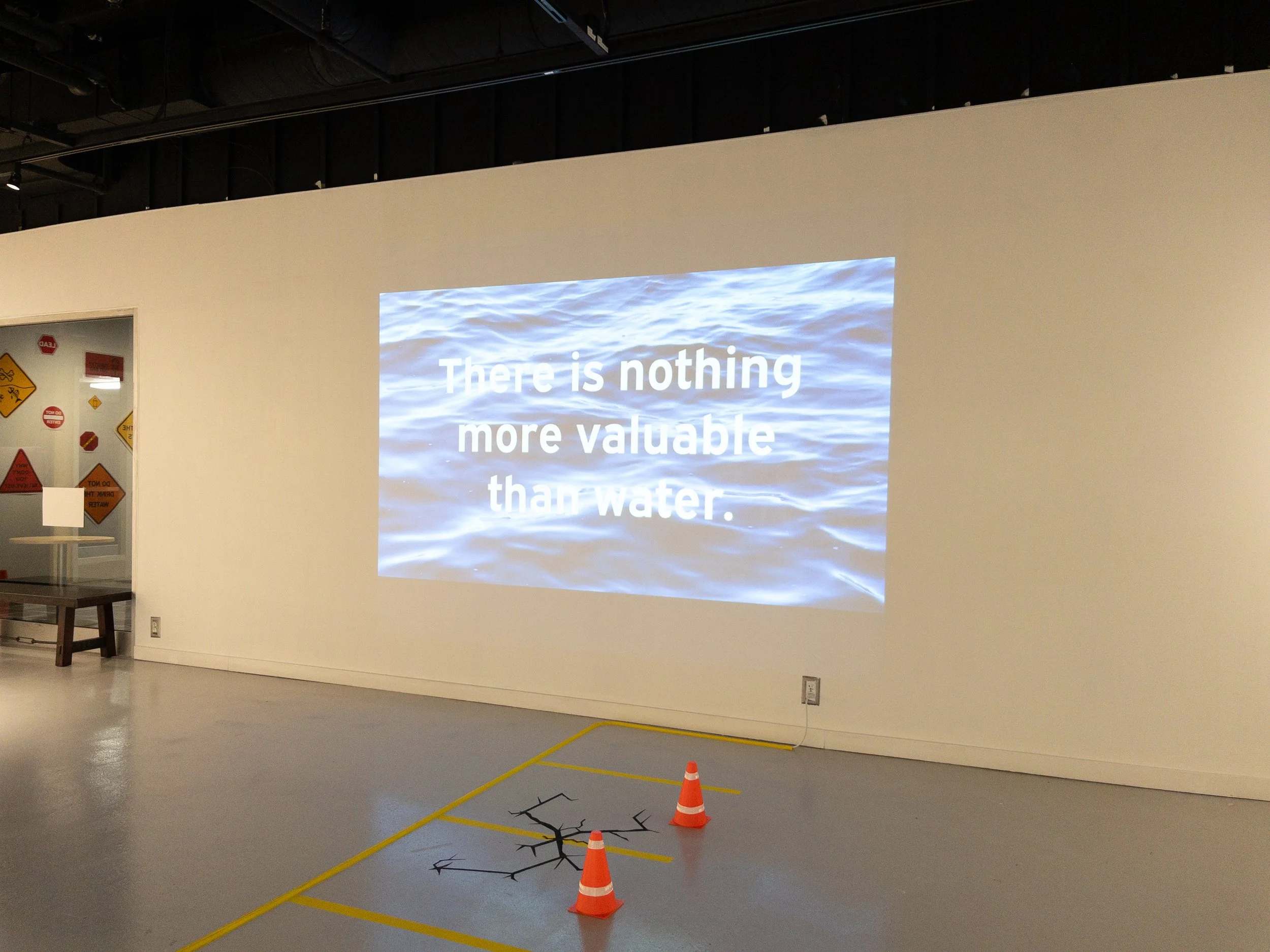 Another section of the exhibition showing a projection, traffic cones, and cracked pavement and parking spots depicted with tape on the floor.