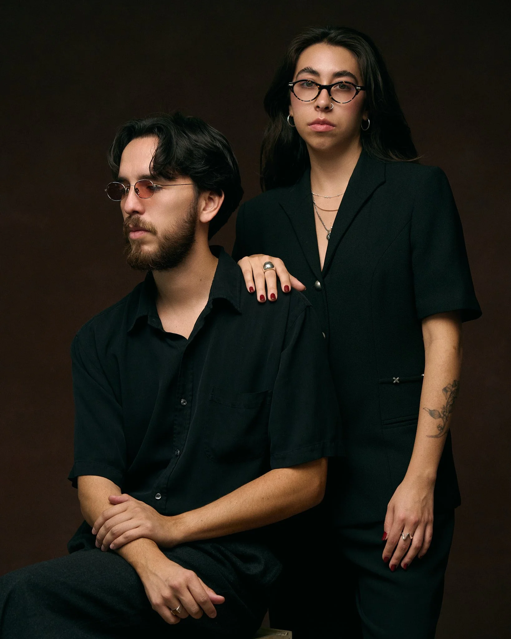 A man with a beard and glasses sitting with his eyes closed, wearing a black shirt; a woman with glasses and tattoo on arm standing next to him with hand on his shoulder, both dressed in black against a dark background.