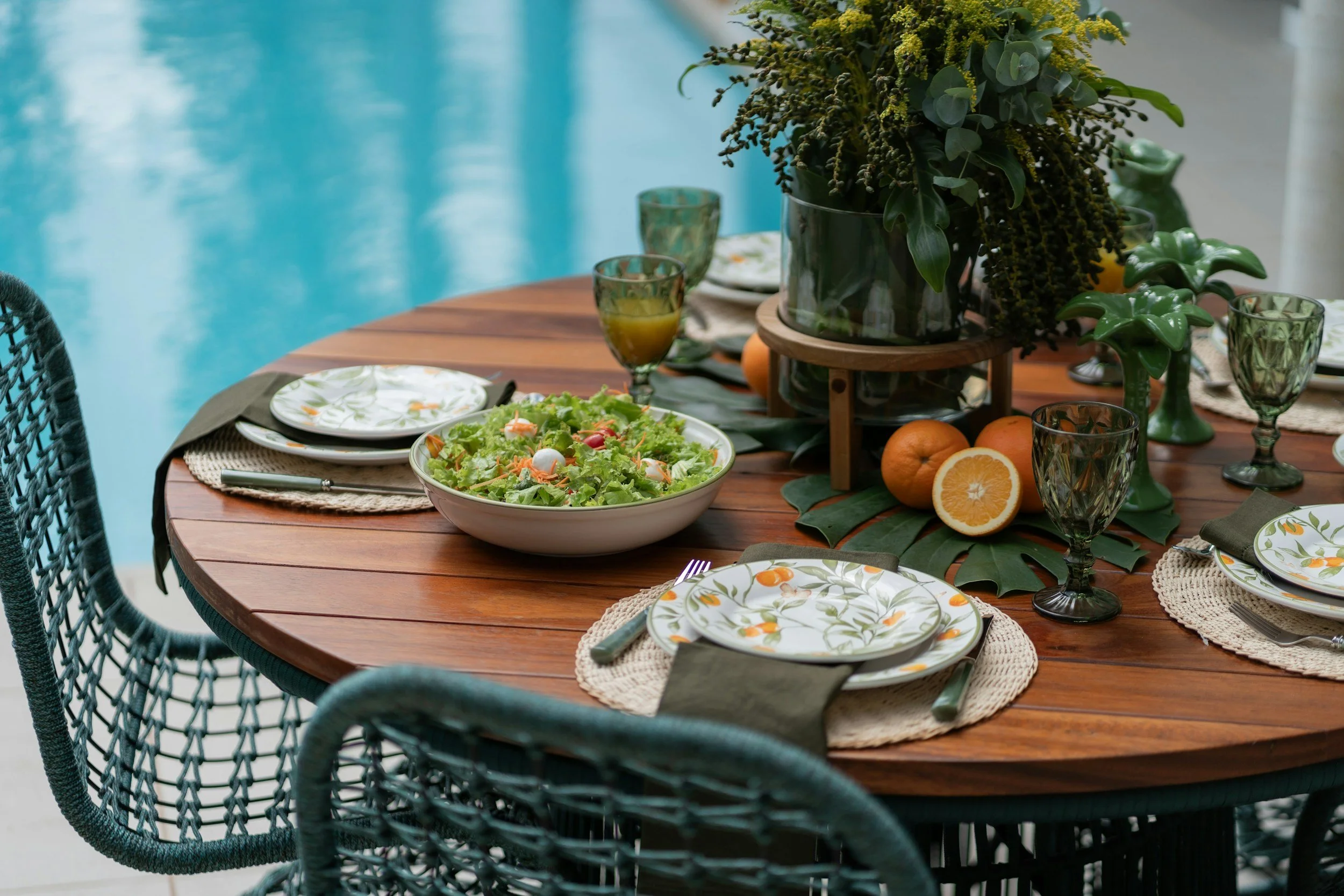 A wooden outdoor dining table by a swimming pool, set with a salad bowl, green patterned plates, glasses of juice, black napkins, and surrounded by dark wicker chairs, with a large floral arrangement in the center.