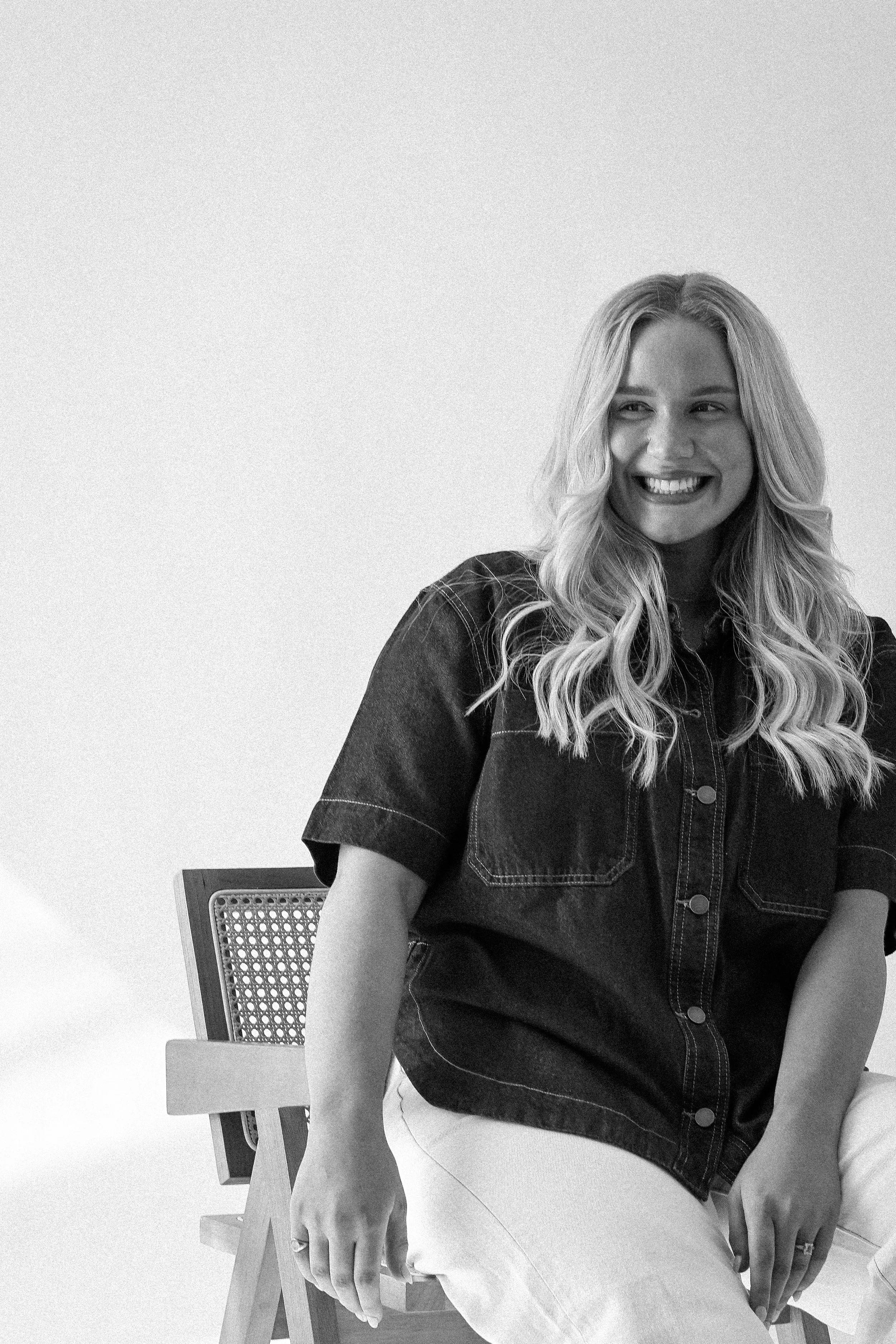 A woman with long wavy hair smiling and sitting on a wooden chair with a cane backrest, wearing a dark short-sleeved denim shirt and light-colored pants.
