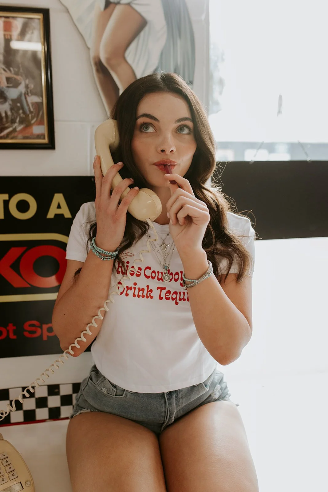 Young woman with dark wavy hair, wearing a white T-shirt and denim shorts, sitting on a table and talking on a vintage beige rotary phone, holding a finger to her lips.