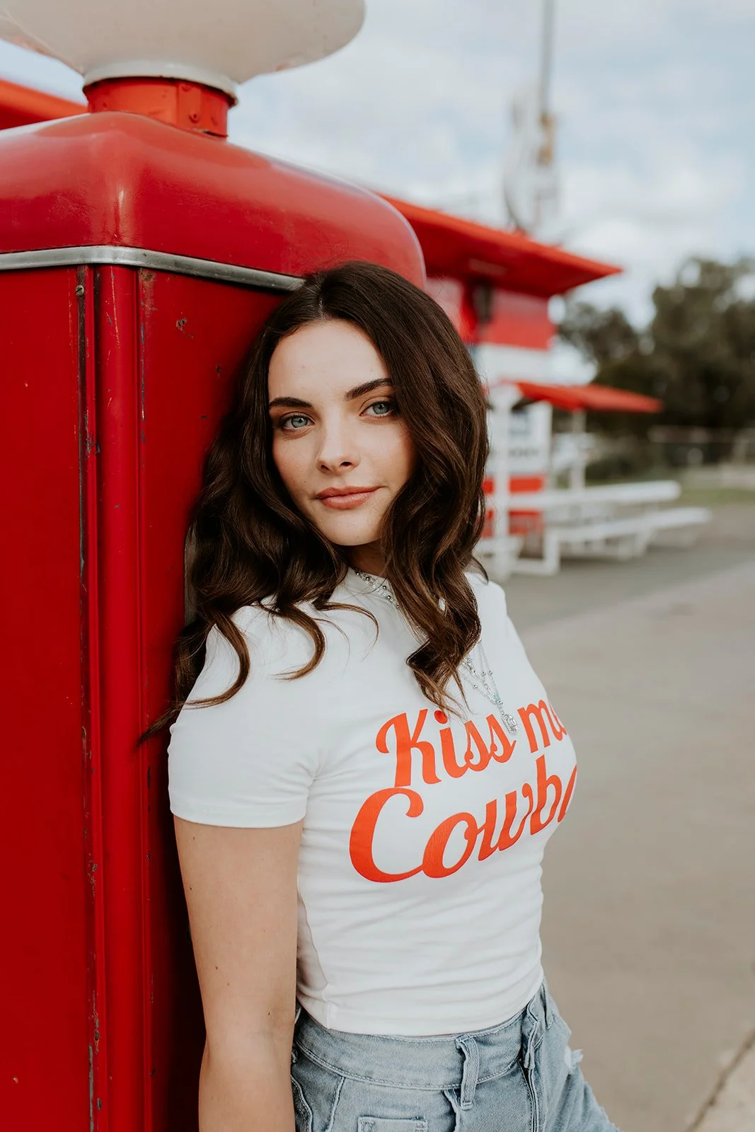 Young woman with long wavy hair leaning against a red vintage gas pump at an outdoor diner or drive-in, wearing a white t-shirt with red lettering and light blue jeans.