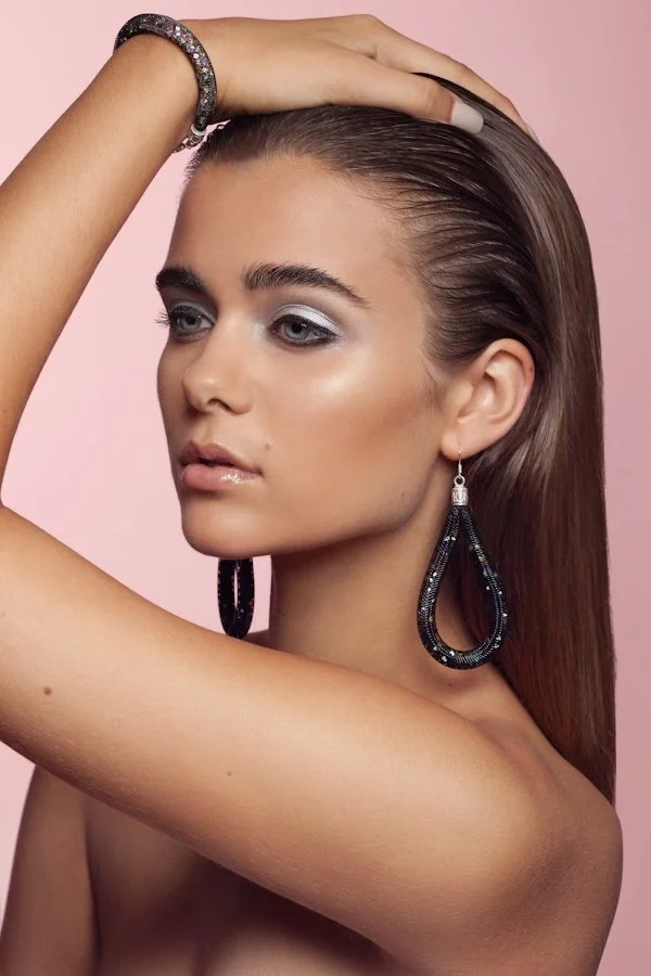 A woman with long brown hair and hoop earrings, posing with her hand on her head, against a pink background.