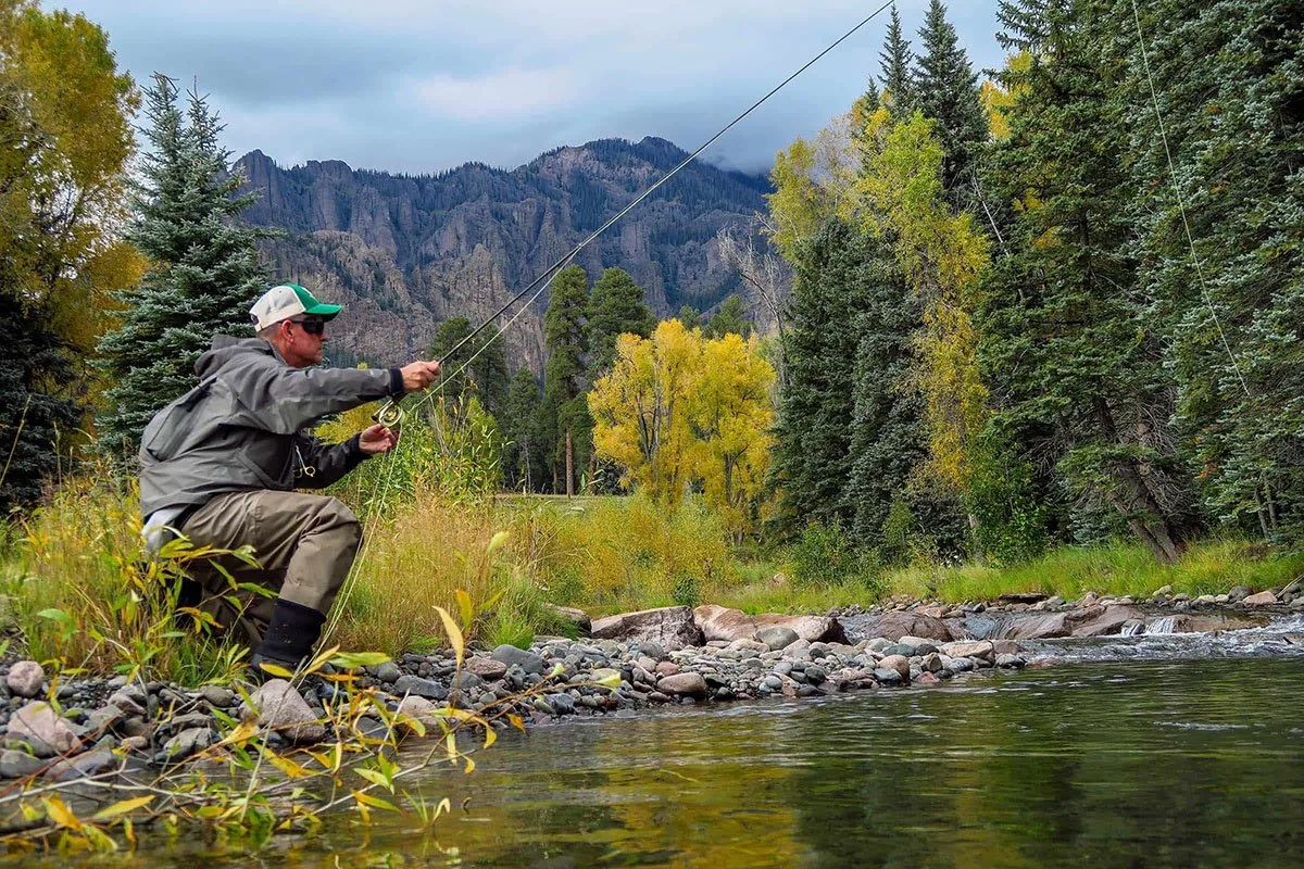 Man fly fishing in a river surrounded by trees and mountains, wearing waders and a cap, with autumn foliage in the background.