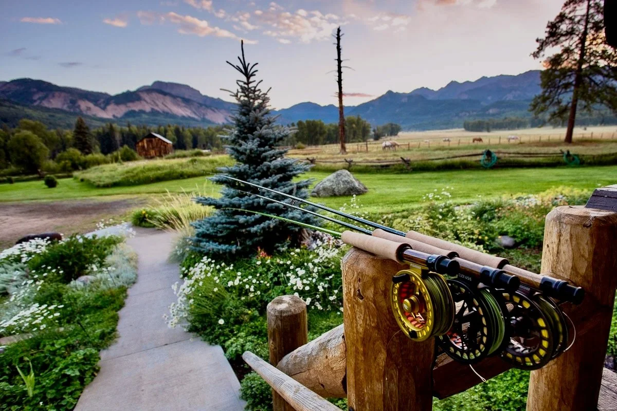 Three fishing rods rest on a wooden fence overlooking a scenic, mountainous landscape with fields, trees, and a cabin at sunset.