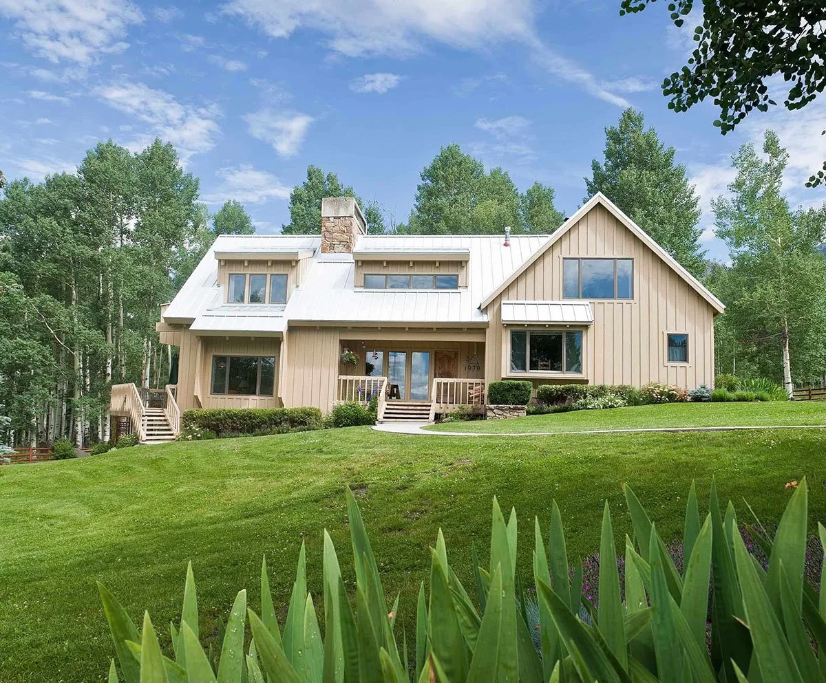 Modern beige house with large windows and a metal roof, surrounded by green grass, trees, and blue sky.
