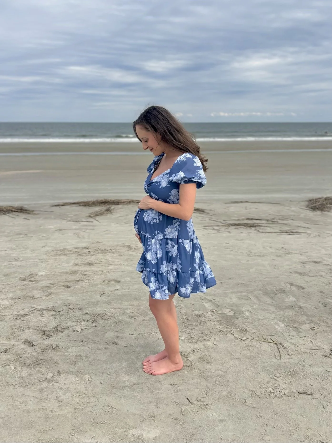 A pregnant woman in a blue floral dress standing barefoot on the sandy beach, looking down and holding her belly with her right hand. Overcast sky and ocean in the background.
