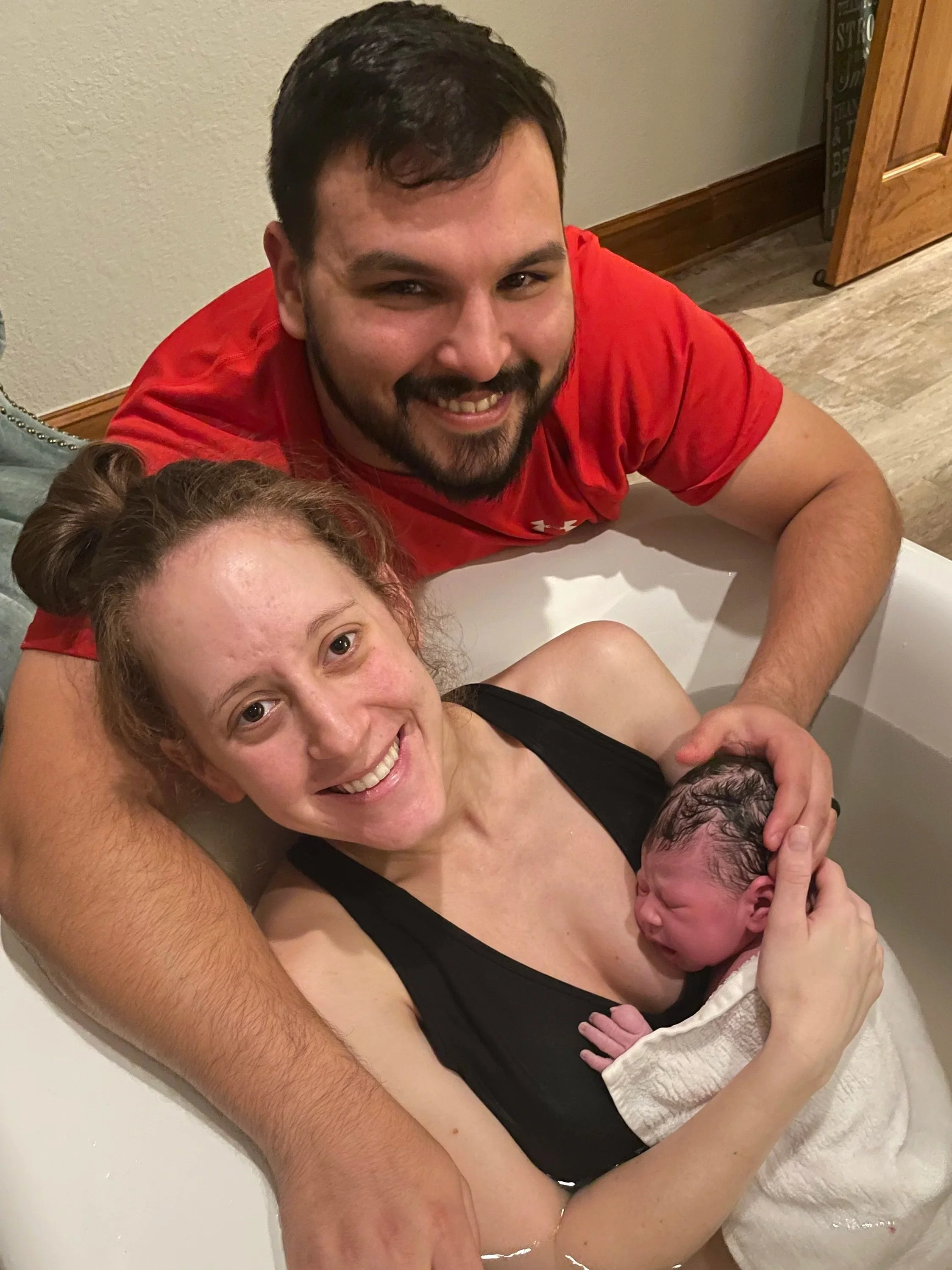 A woman with red curly hair in a black tank top holding a newborn in a towel, with a smiling man in a red shirt leaning over the bathtub with his arm around her.