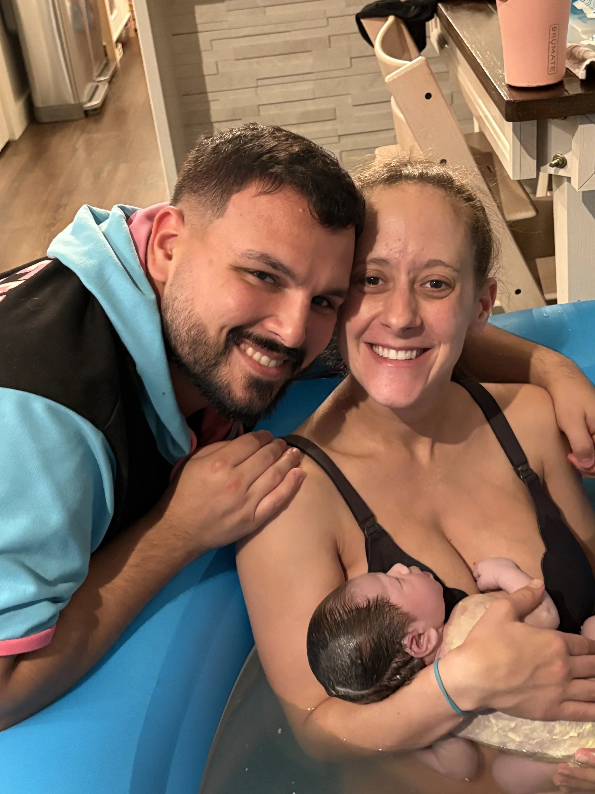 A woman holding a newborn baby in a bath, with a man leaning over and smiling beside them, inside a home.