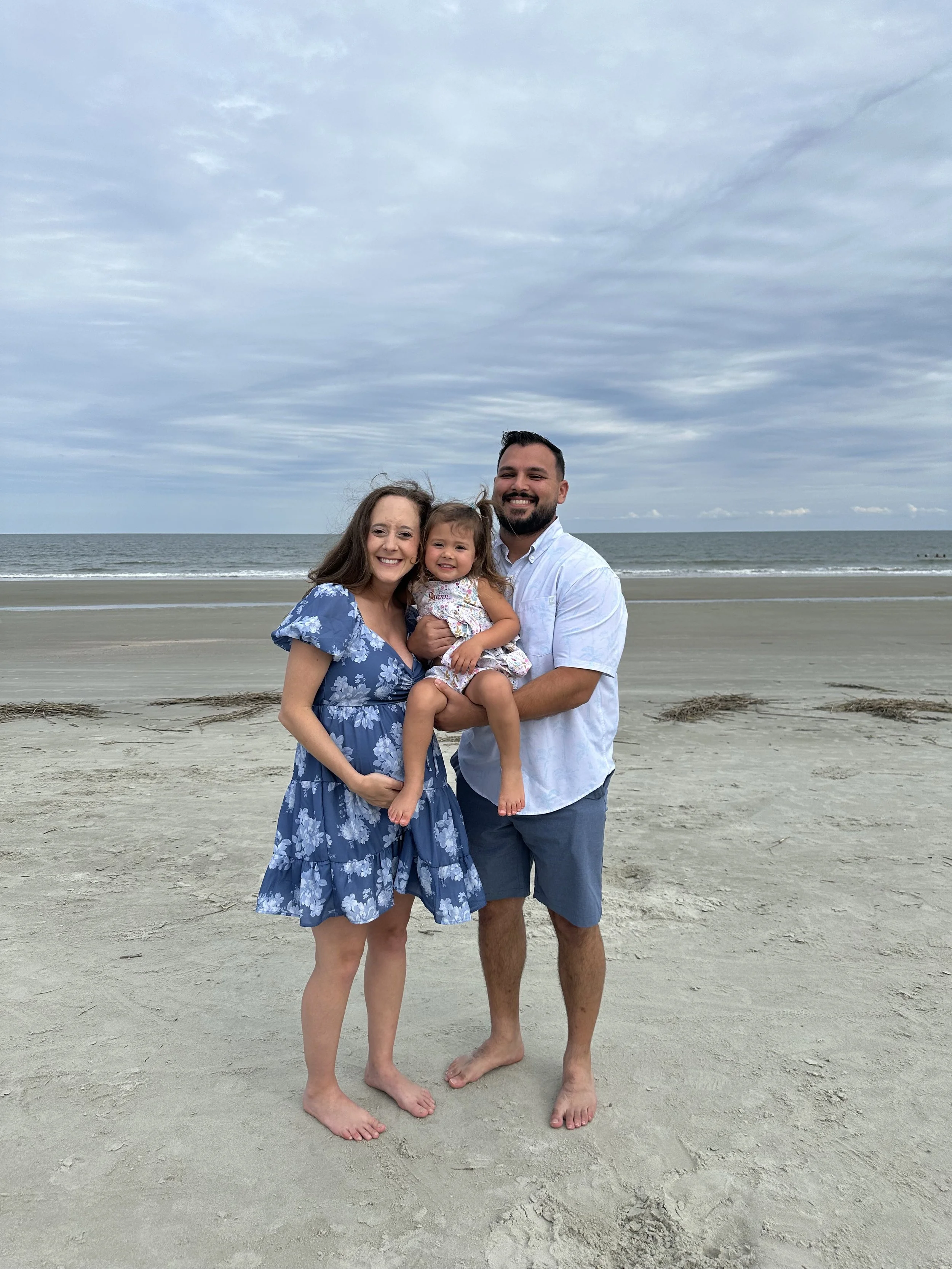 Family of three smiling, standing on a sandy beach near the ocean, under a cloudy sky, with the woman in a blue floral dress, the man in a white shirt and blue shorts, and a young girl in a floral dress.