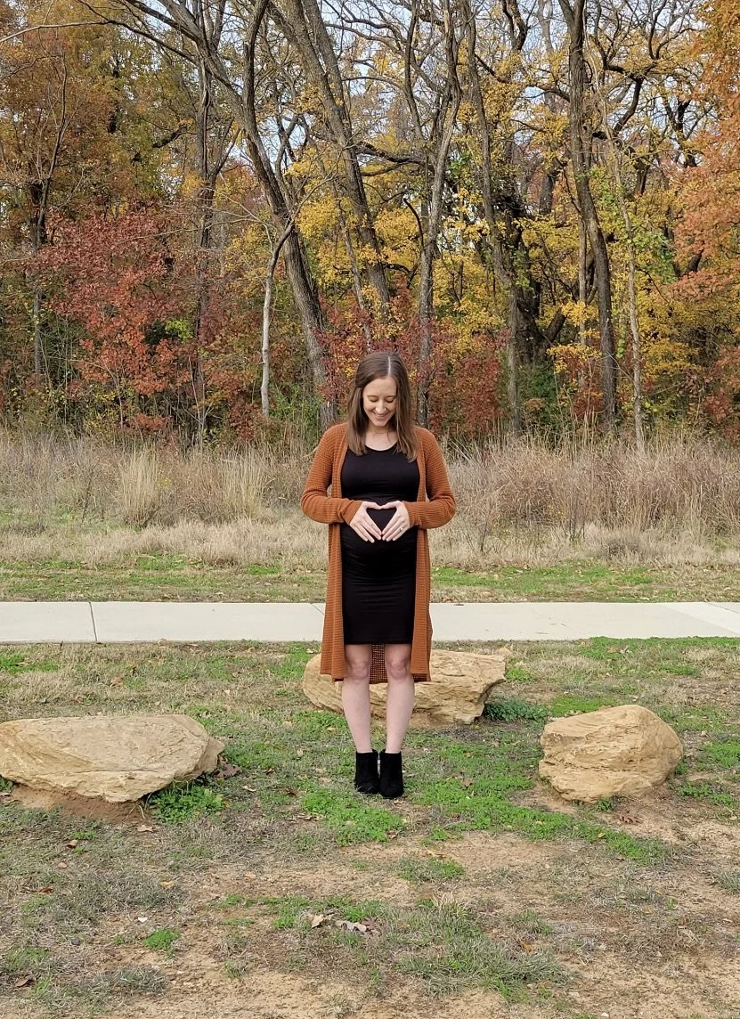 A pregnant woman smiling and forming a heart with her hands on her belly, standing outdoors in front of autumn-colored trees and large rocks.