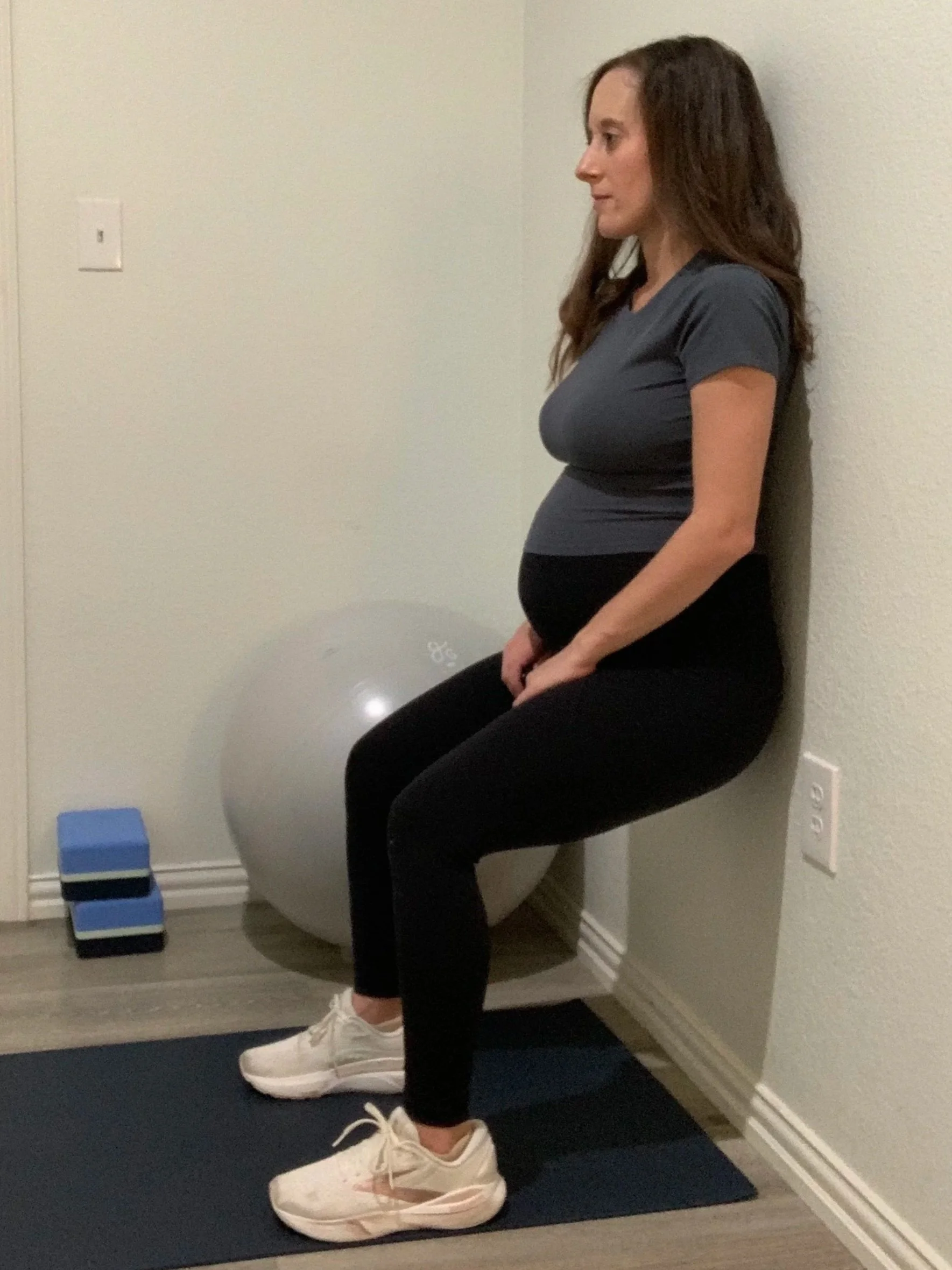 A pregnant woman sitting against a wall in a home gym, with exercise equipment like a stability ball, blue foam blocks, and a yoga mat visible around her.