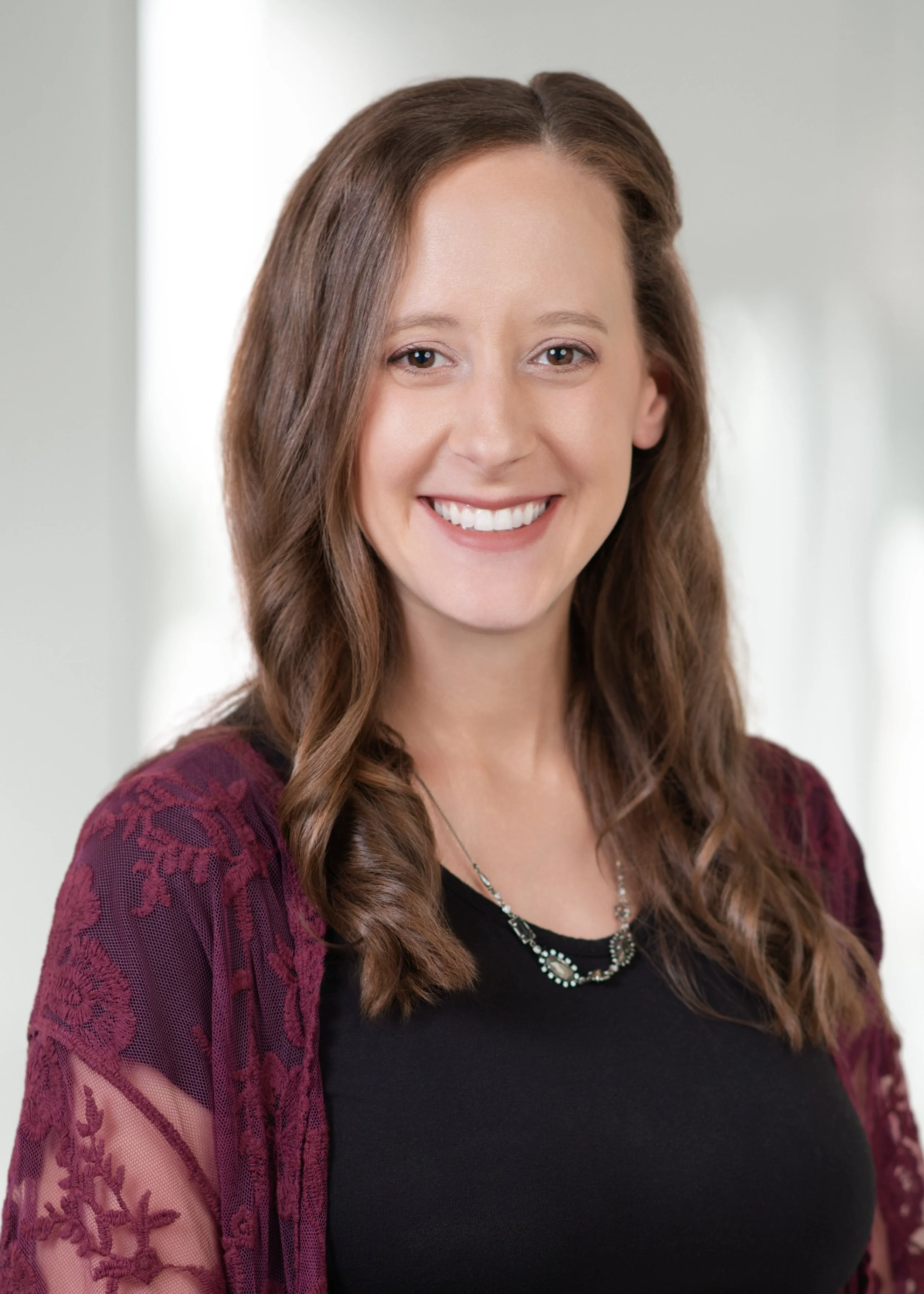 A woman with long, wavy brown hair smiling, wearing a black top and a maroon lace cardigan, jewelry, in a bright indoor setting.