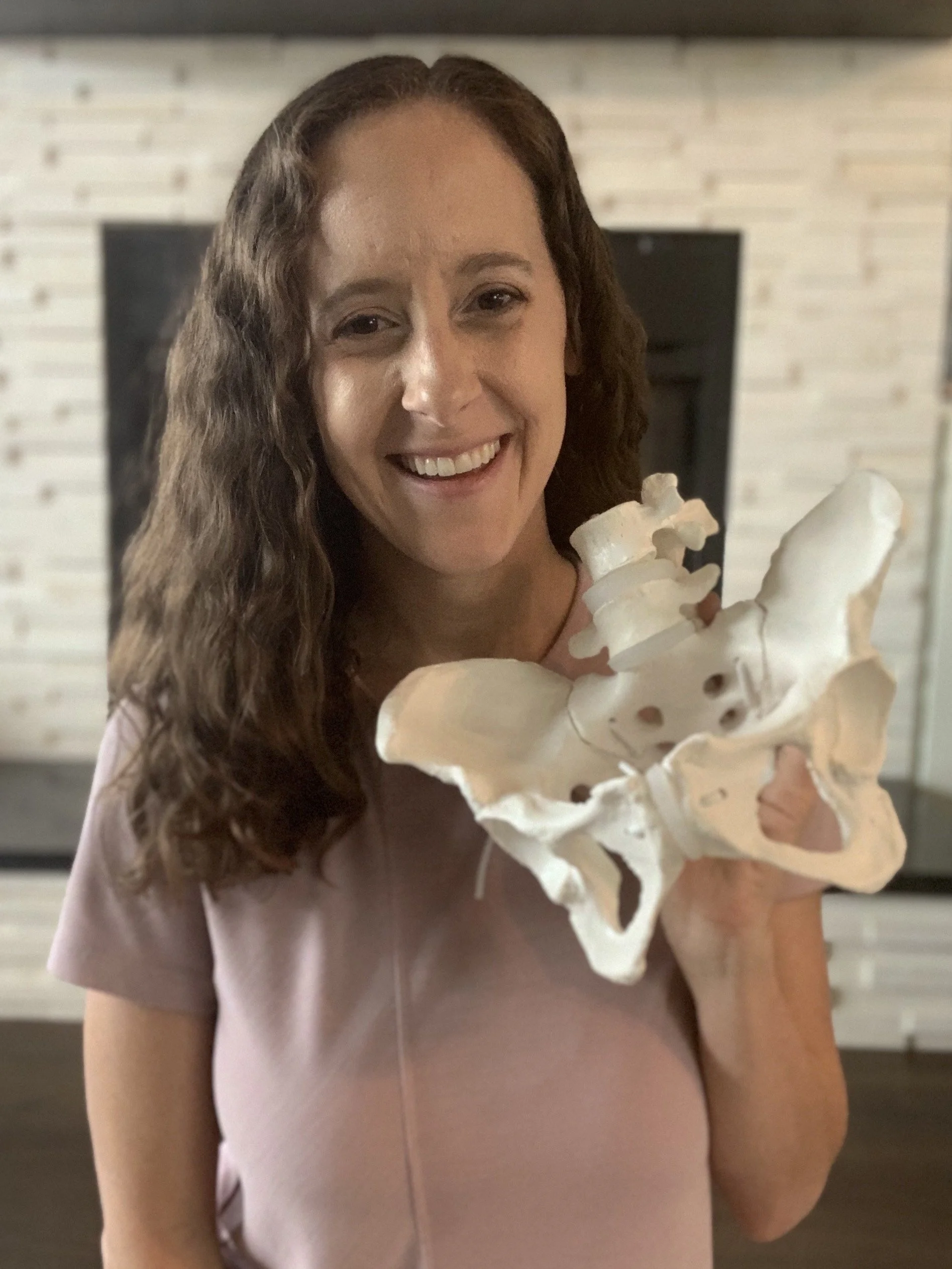 A woman with curly hair smiling and holding a large model of a human pelvis with lumbar vertebrae.