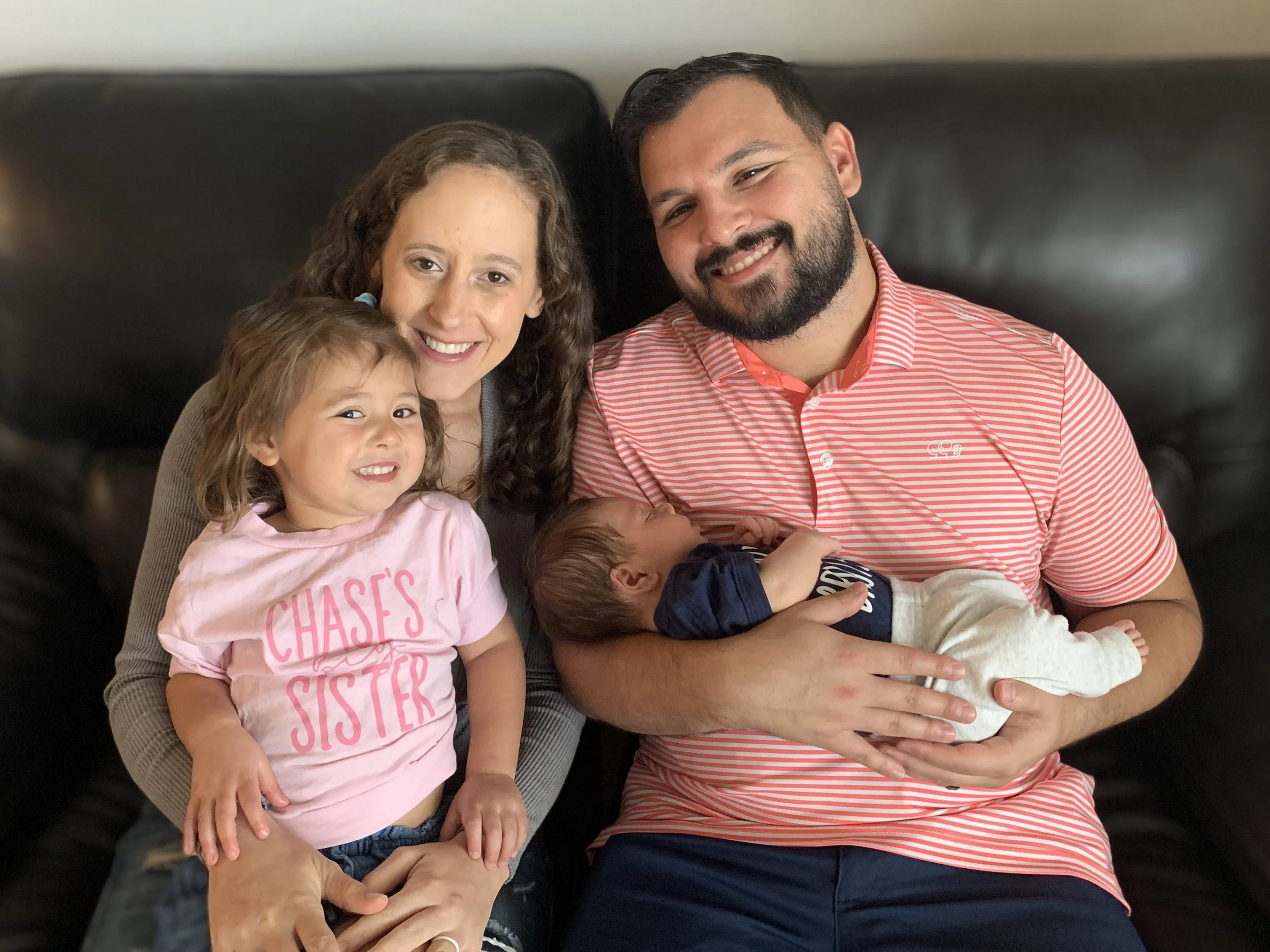 A family of four sitting on a black leather couch, including a woman with curly hair, a man with a beard, a young girl smiling, and a newborn baby being held by the man.