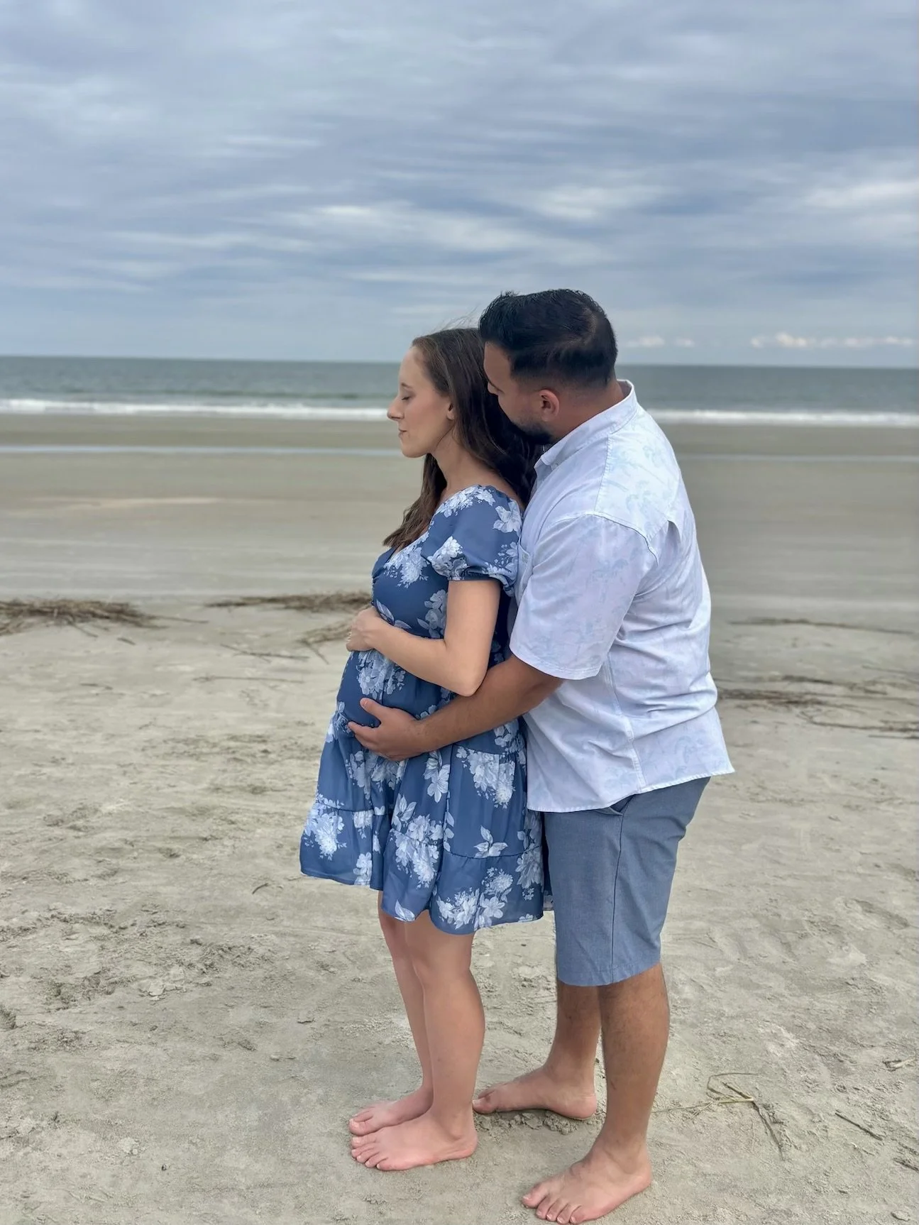 A couple stands on a beach embracing each other, with the ocean and cloudy sky in the background.