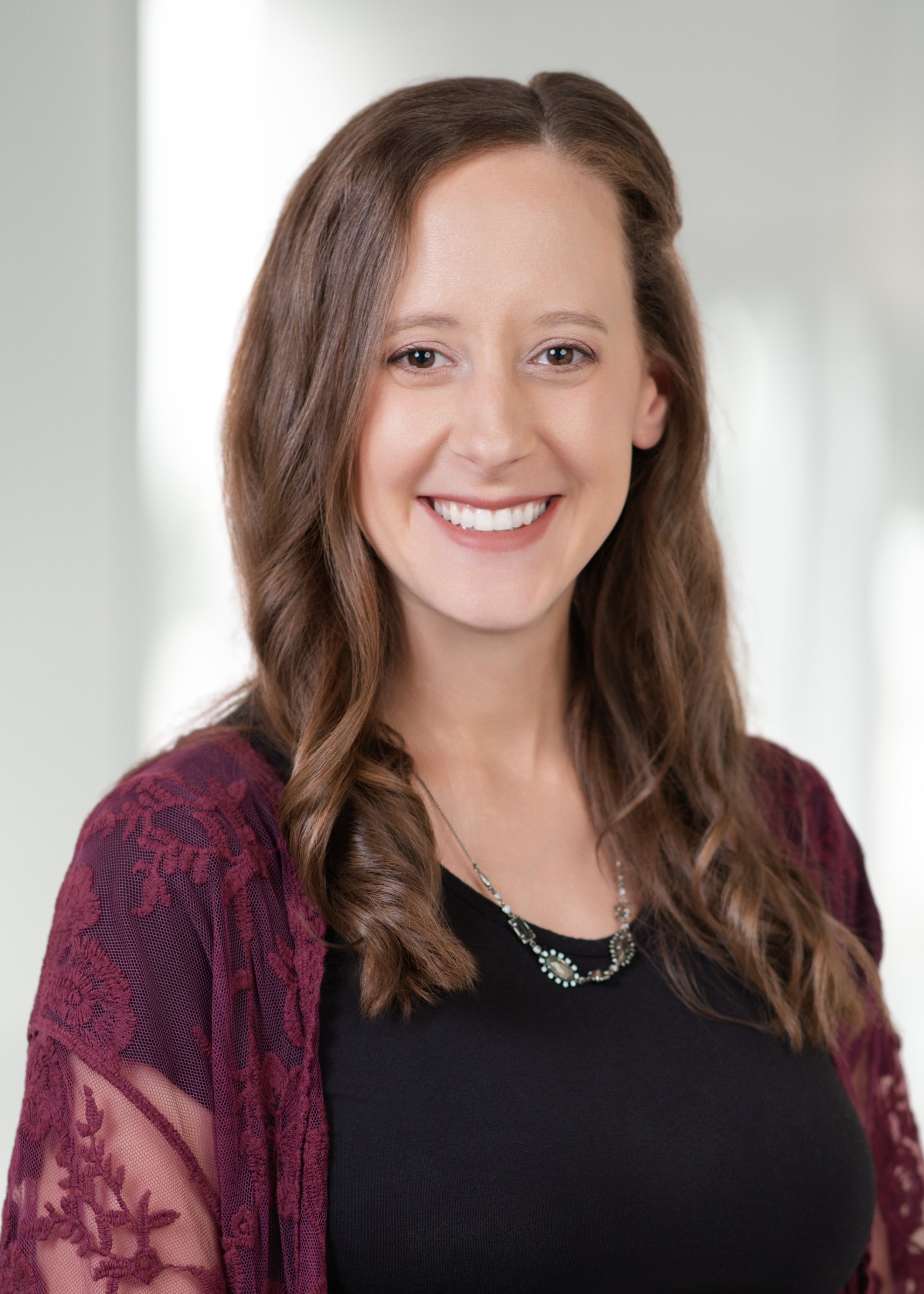 Headshot of a woman with long brown hair smiling, wearing a black top and a burgundy lace cardigan, against a blurred indoor background.