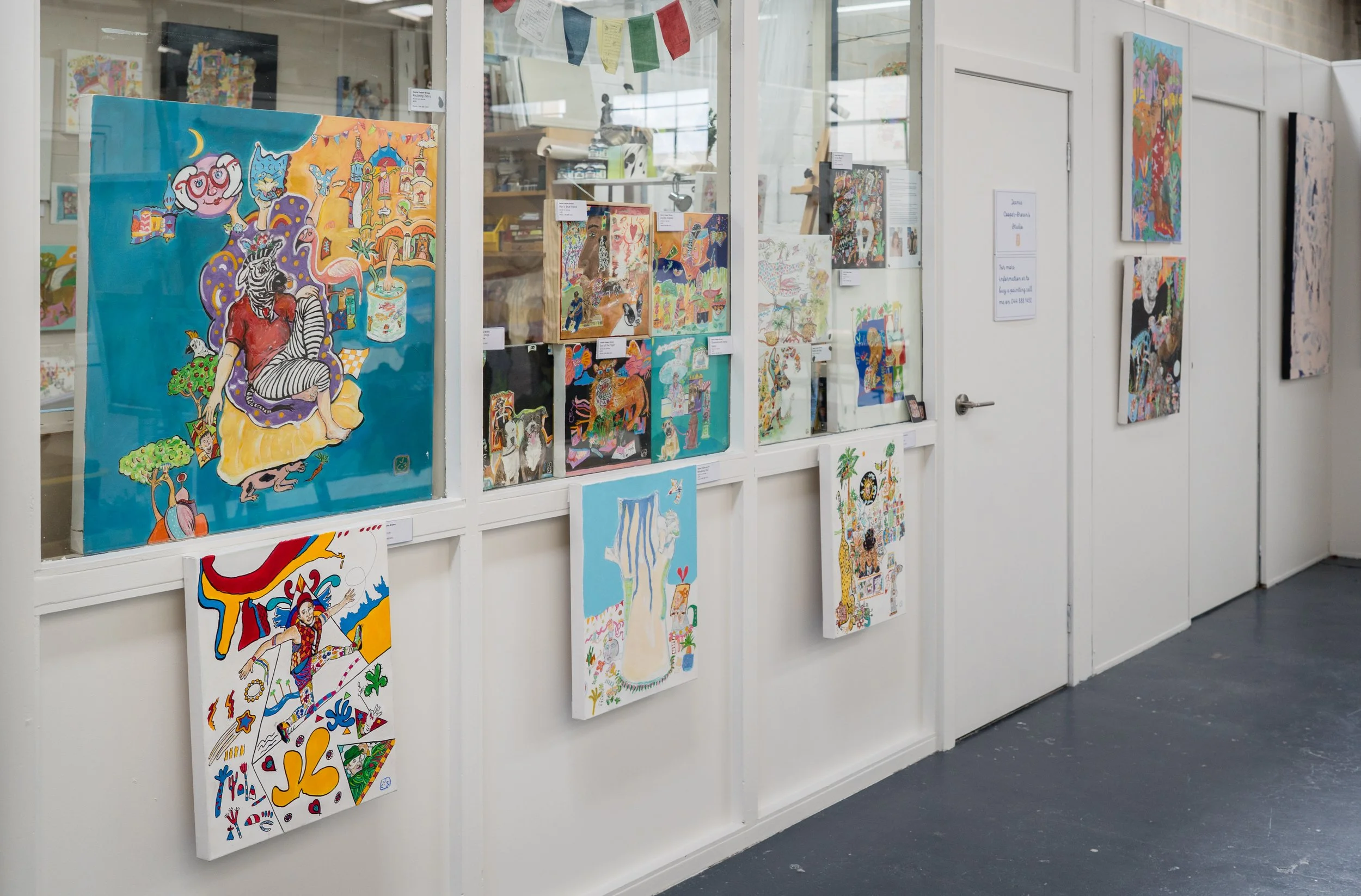 Art gallery display with colorful paintings and drawings on white walls and glass partition.