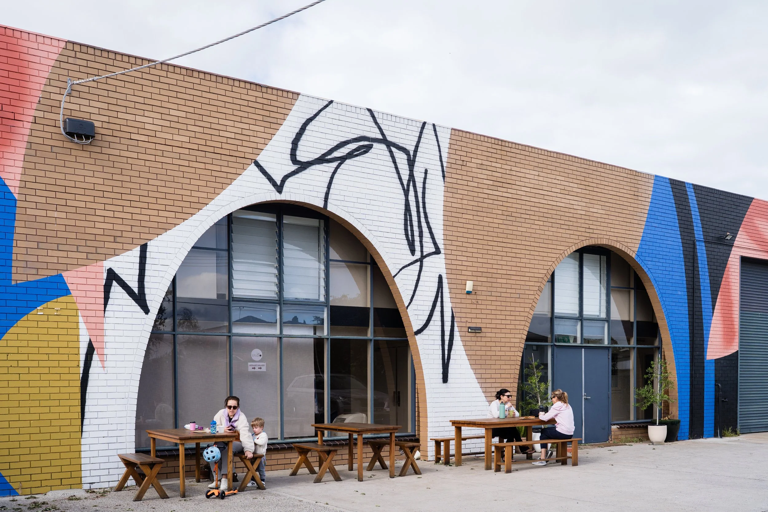 People sitting at outdoor tables in front of a building with colorful abstract mural and large arched windows.