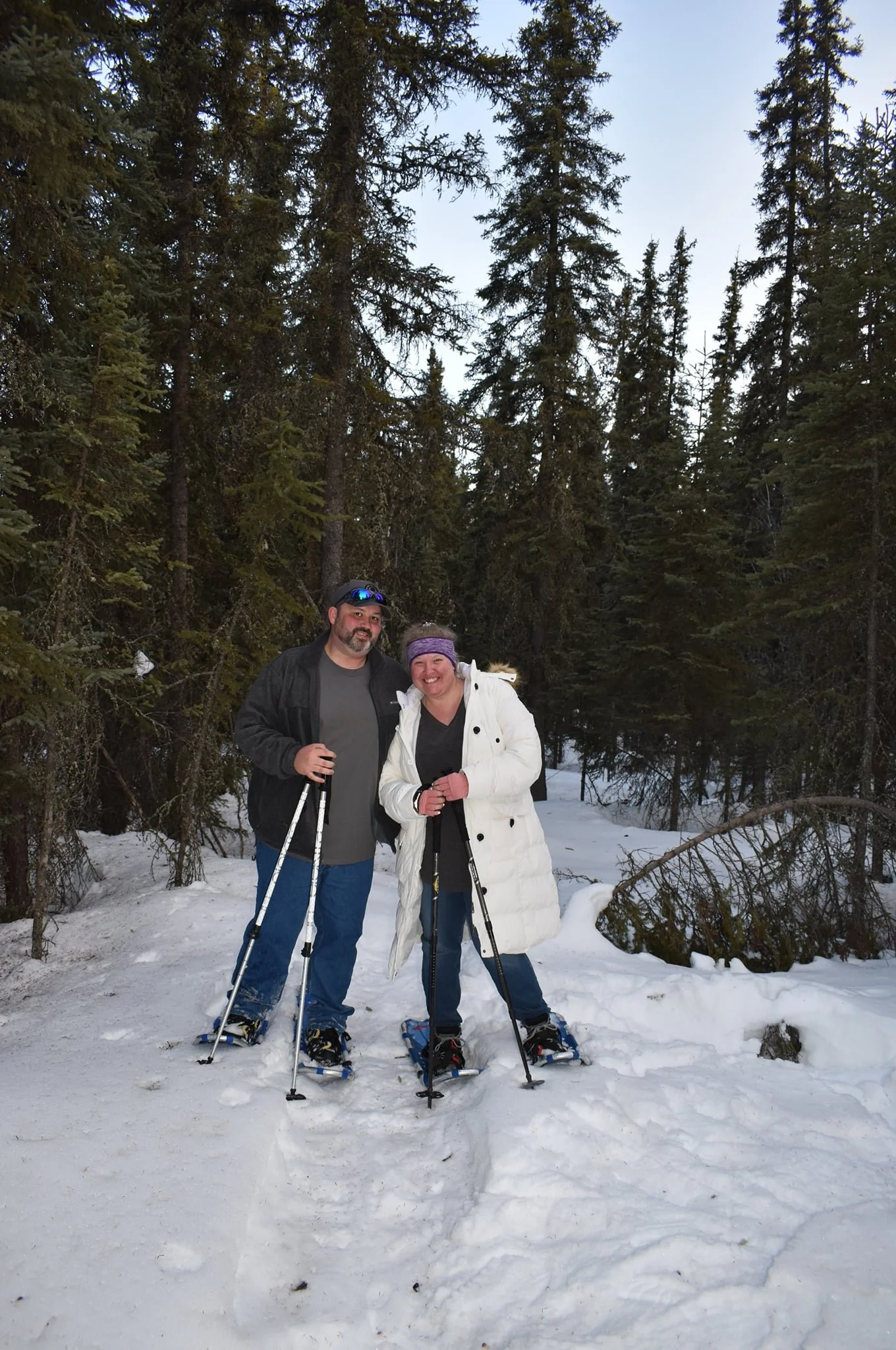 A man and woman on snowshoes standing in a snowy forest, smiling, with tall evergreen trees in the background.