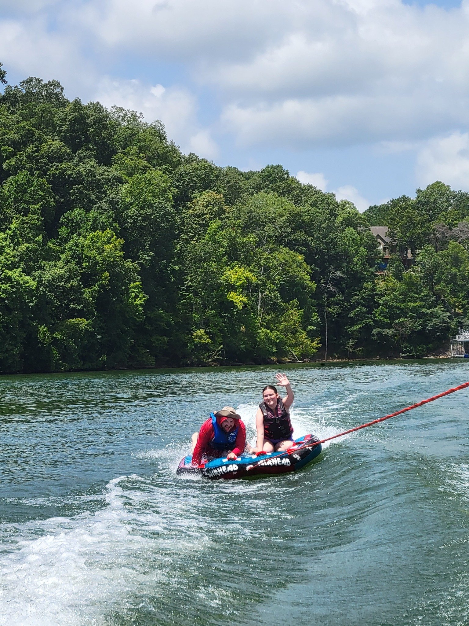 Two people on a wakeboard being pulled by a boat on a lake, waving and smiling, with green trees and a cloudy sky in the background.