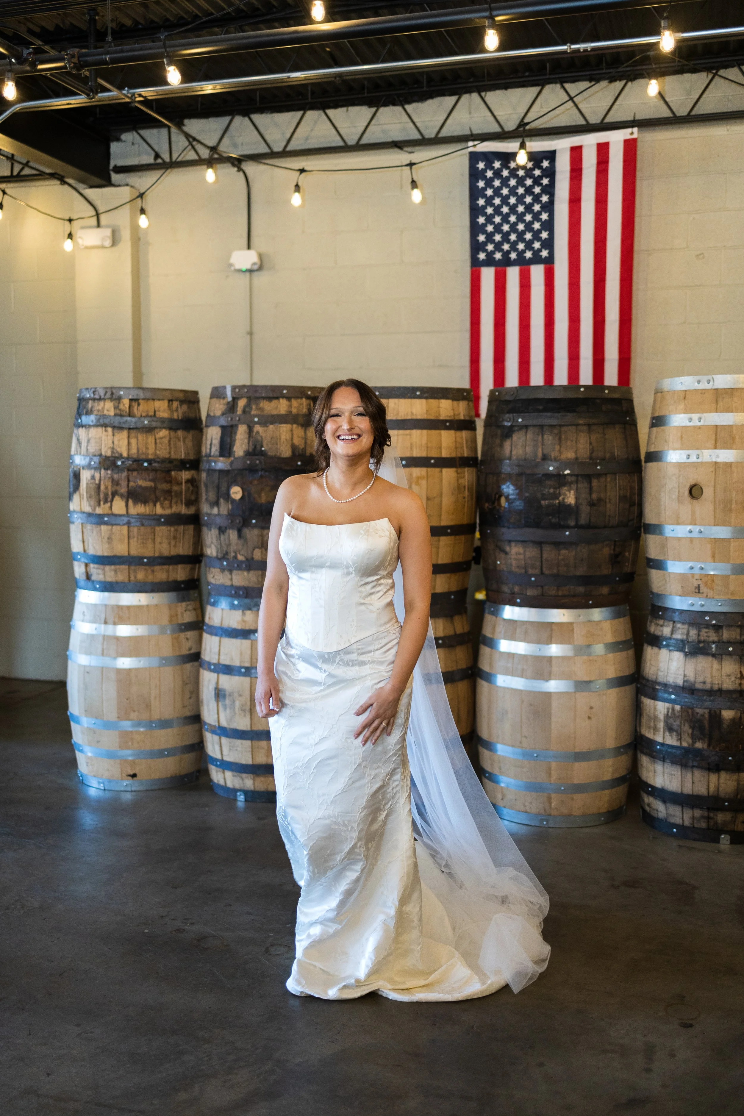 A bride in a white wedding dress smiling indoors, standing in front of stacked whiskey barrels with an American flag hanging on the wall behind her.