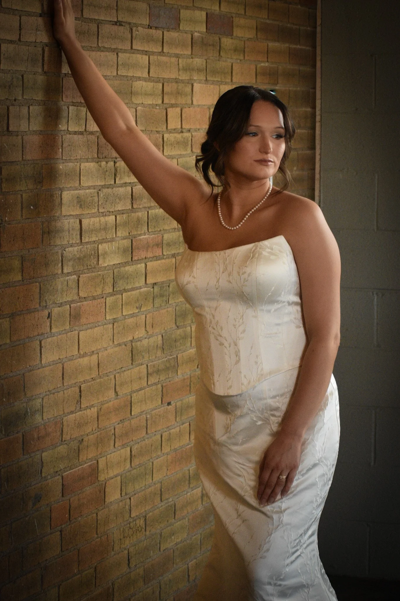 A woman in a strapless cream-colored dress with a pearl necklace poses against a brick wall.