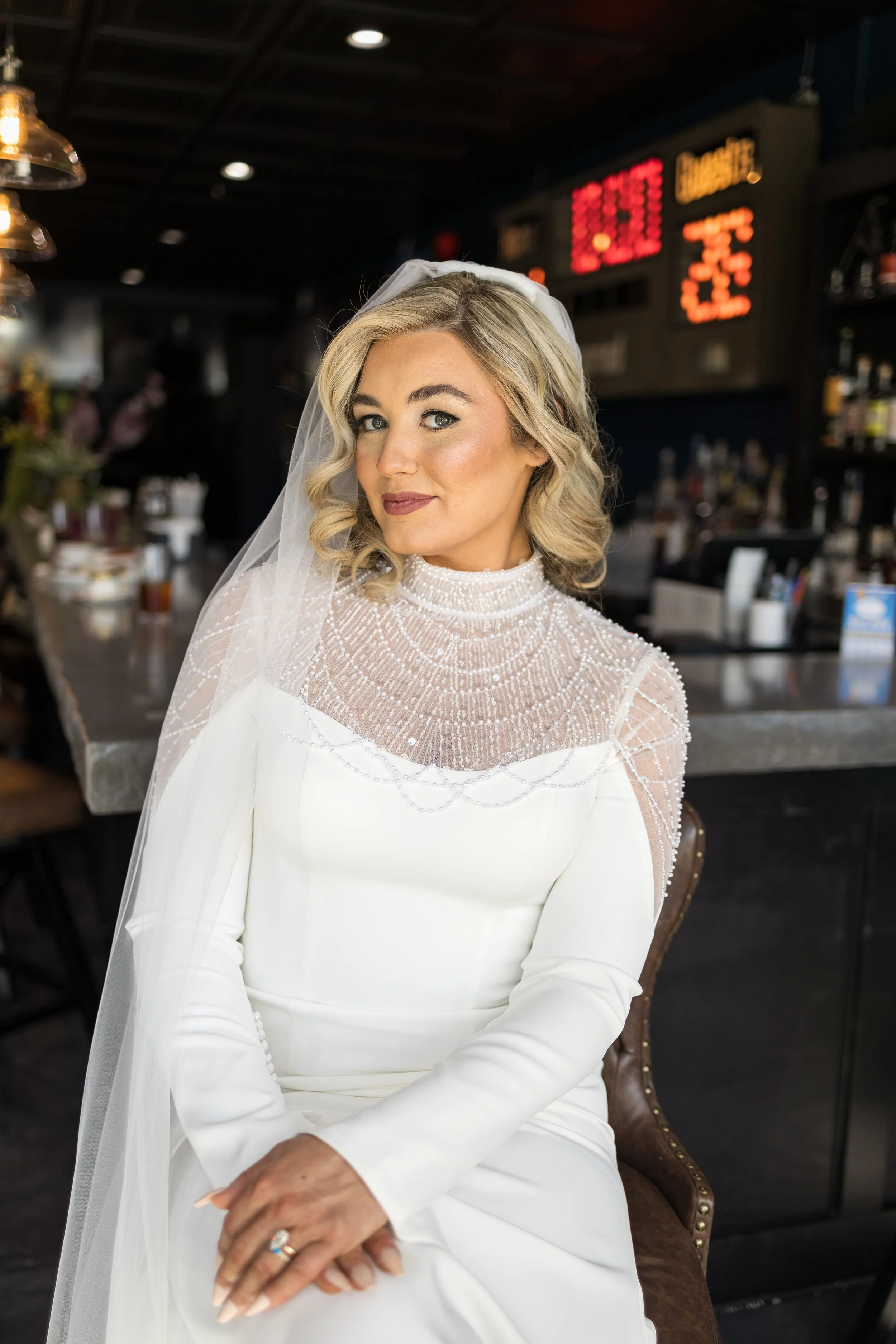 A woman dressed in a white wedding gown with a beaded neckline and a veil sits in a bar or restaurant, looking at the camera.