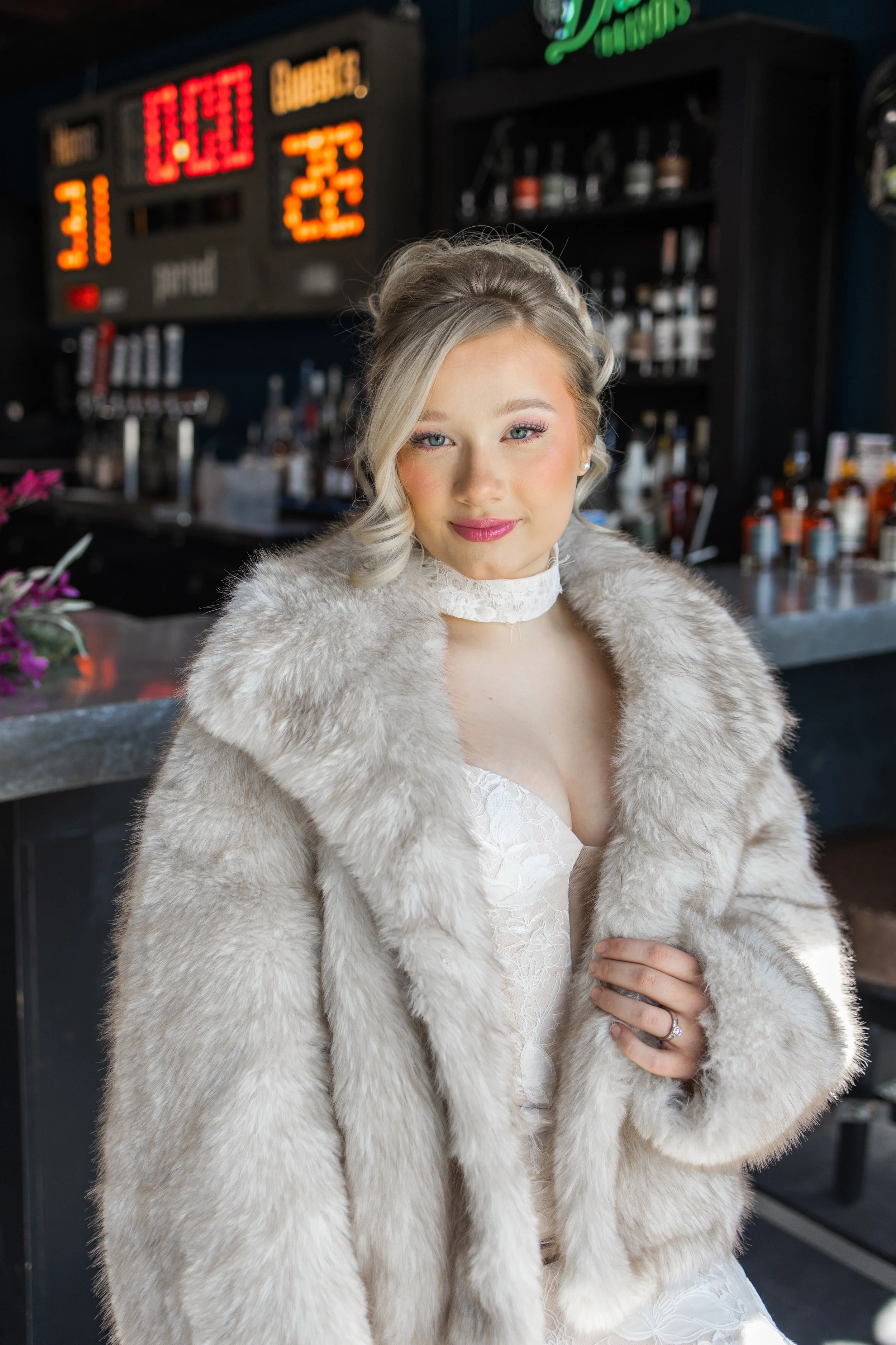 Young woman in a white lace dress and fur coat standing at a bar, with a digital sports scoreboard in the background.