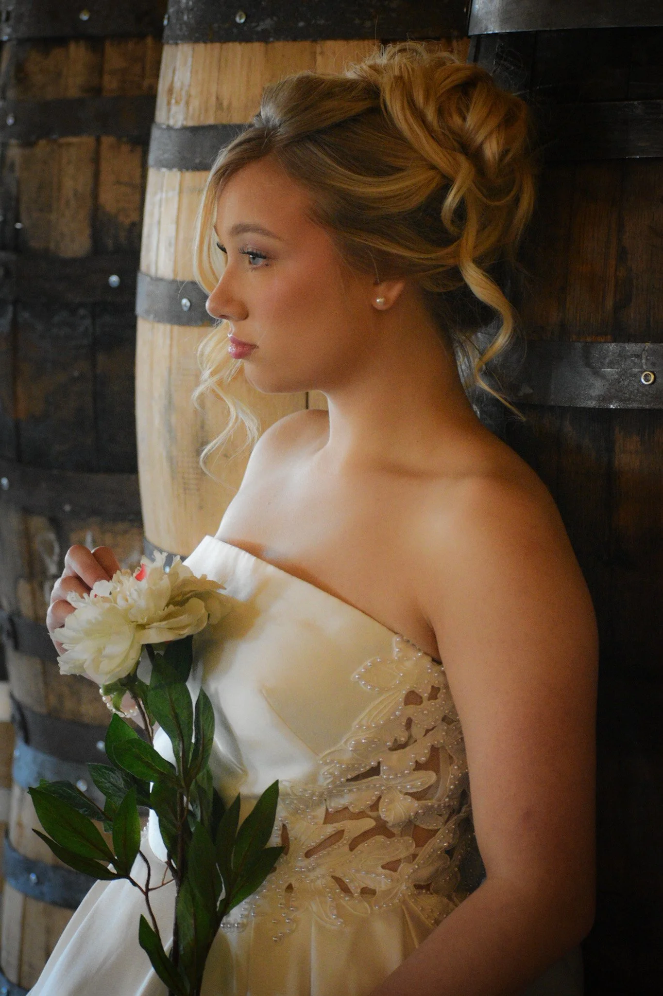 A young woman with blonde hair styled in loose curls, wearing a strapless white dress with intricate lace and bead details, holding a white flower and green leaves, standing against a wooden barrel wall.