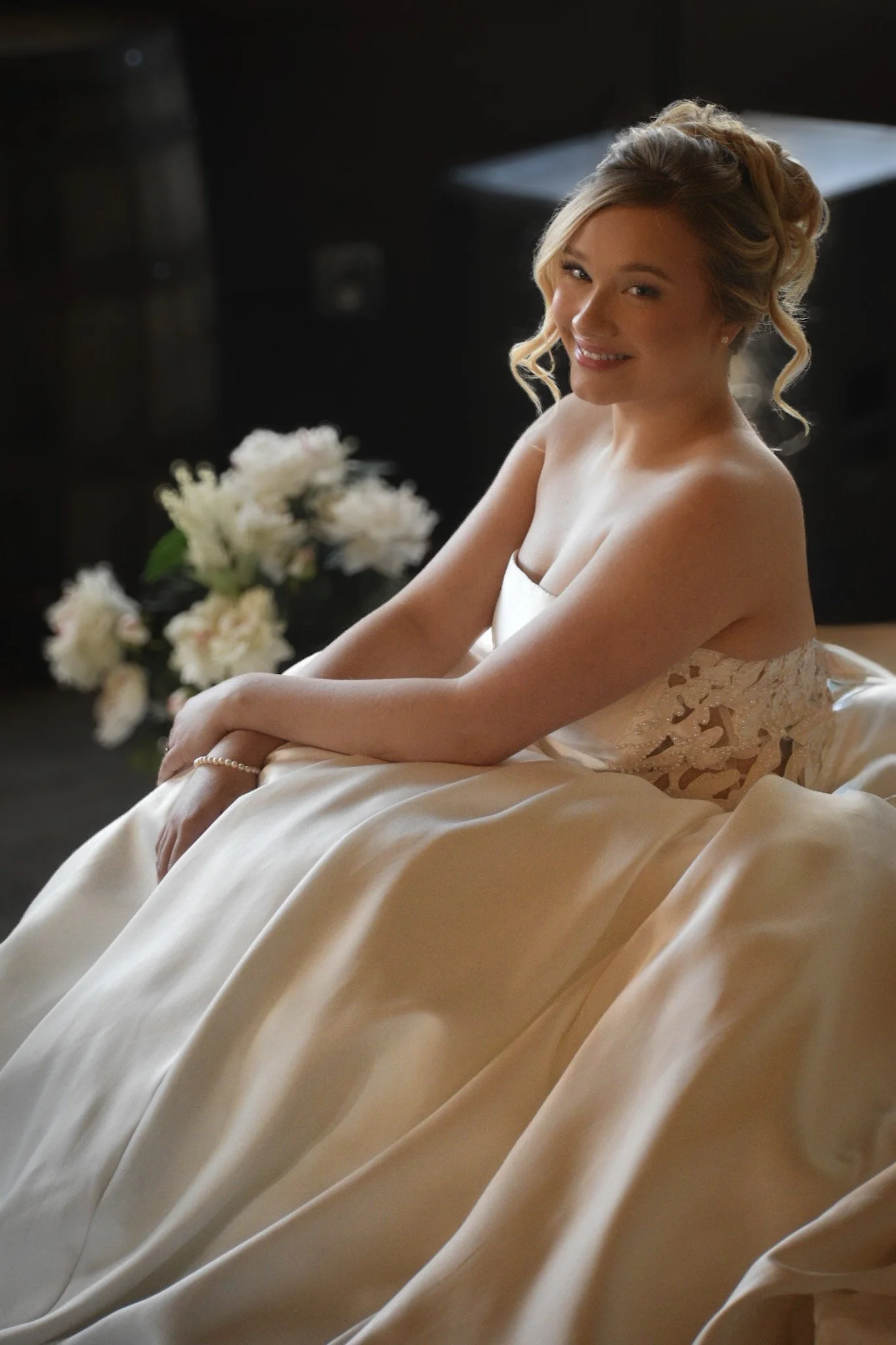 A young woman in a wedding dress sitting on the floor, smiling at the camera with flowers in the background.