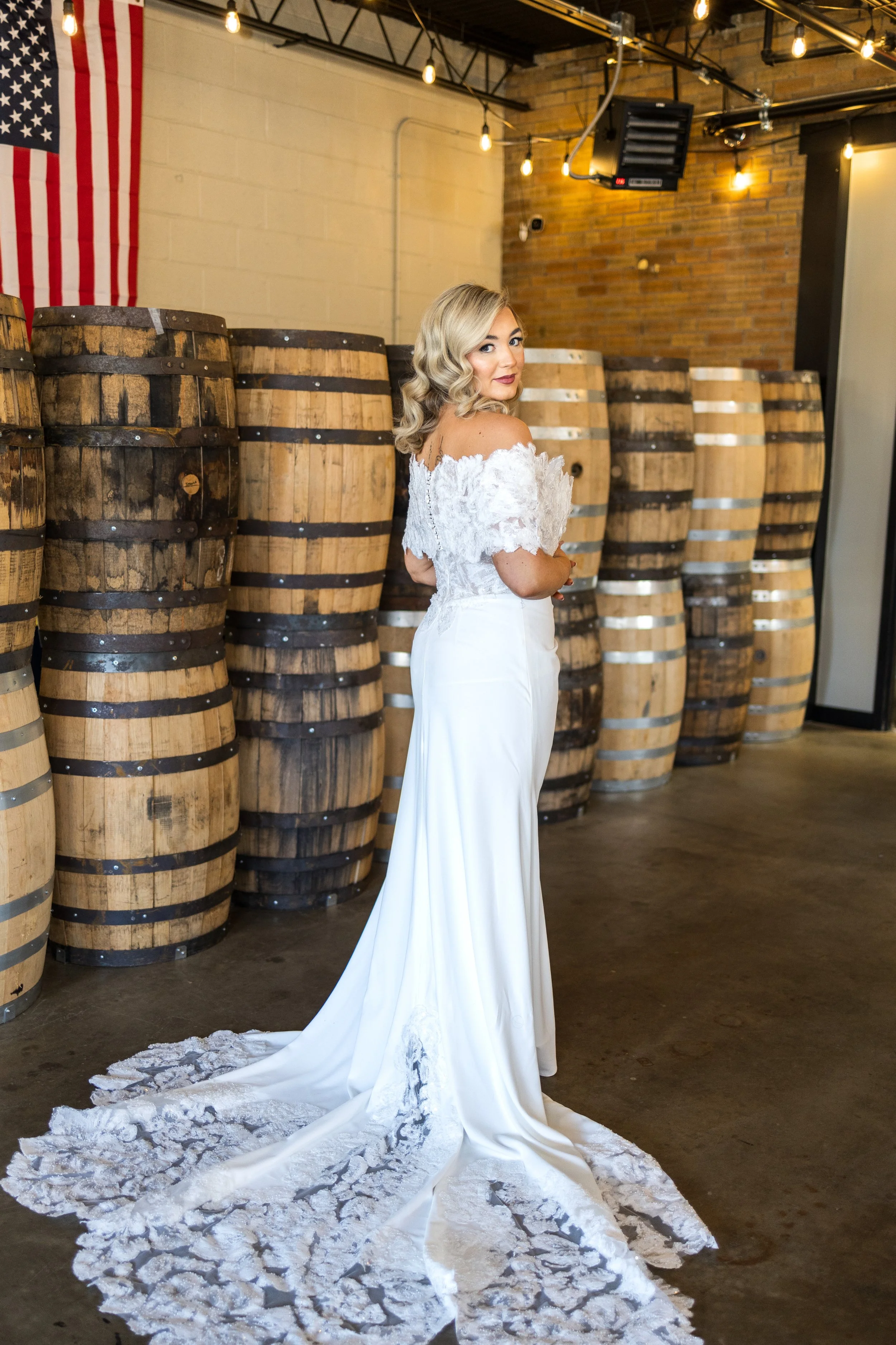 A woman in a white wedding dress with lace details and a long train, standing indoors with barrels in the background and an American flag on the wall.