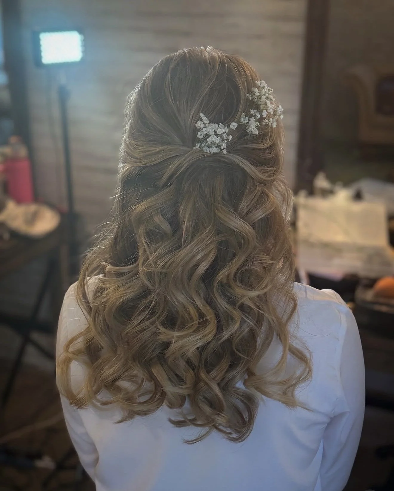 Back view of woman with wavy blonde hair decorated with baby's breath flowers, wearing a white garment.