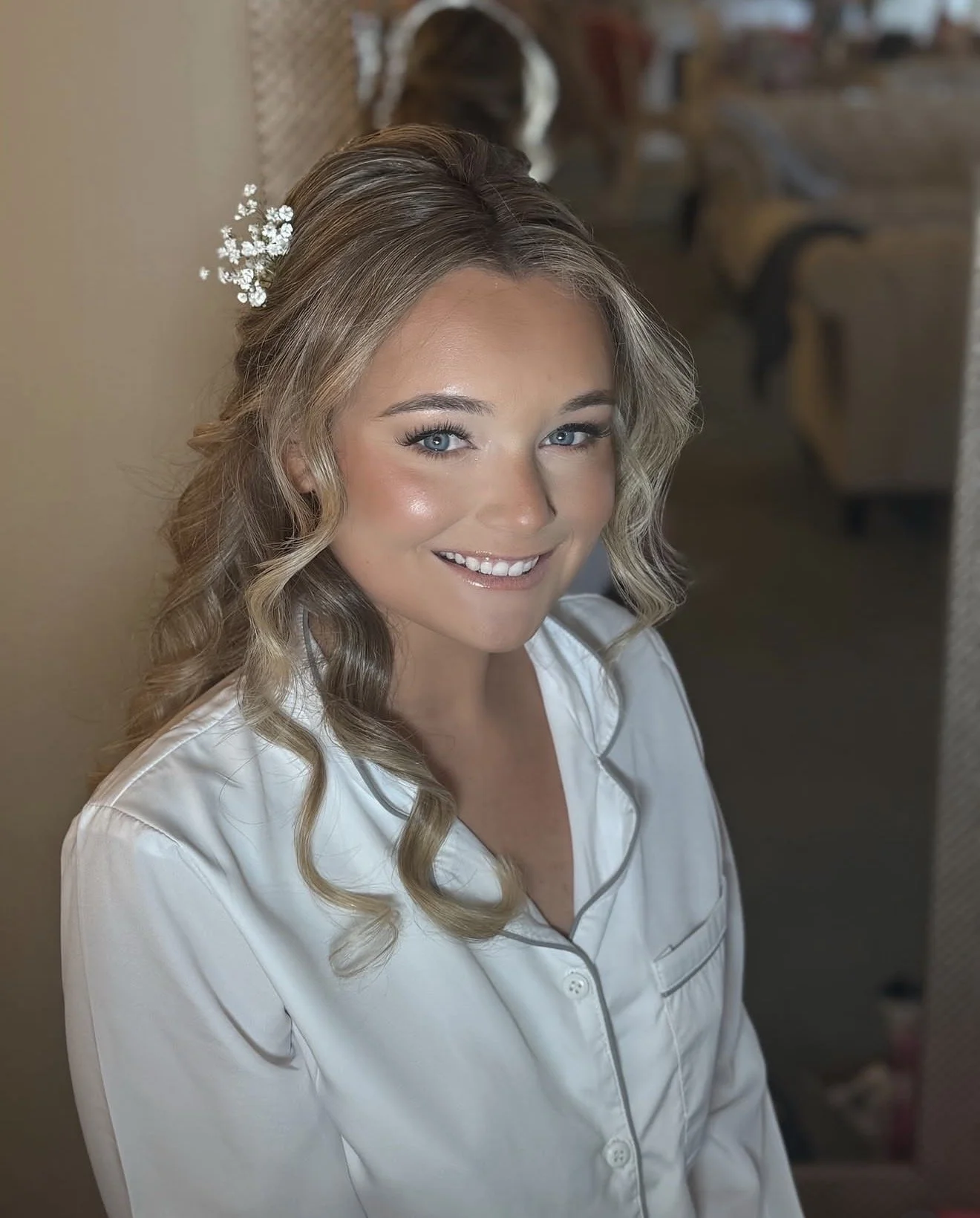 Close-up of a smiling woman with curly blonde hair, wearing a white pajama top, and a white floral hair accessory, in a cozy indoor setting.