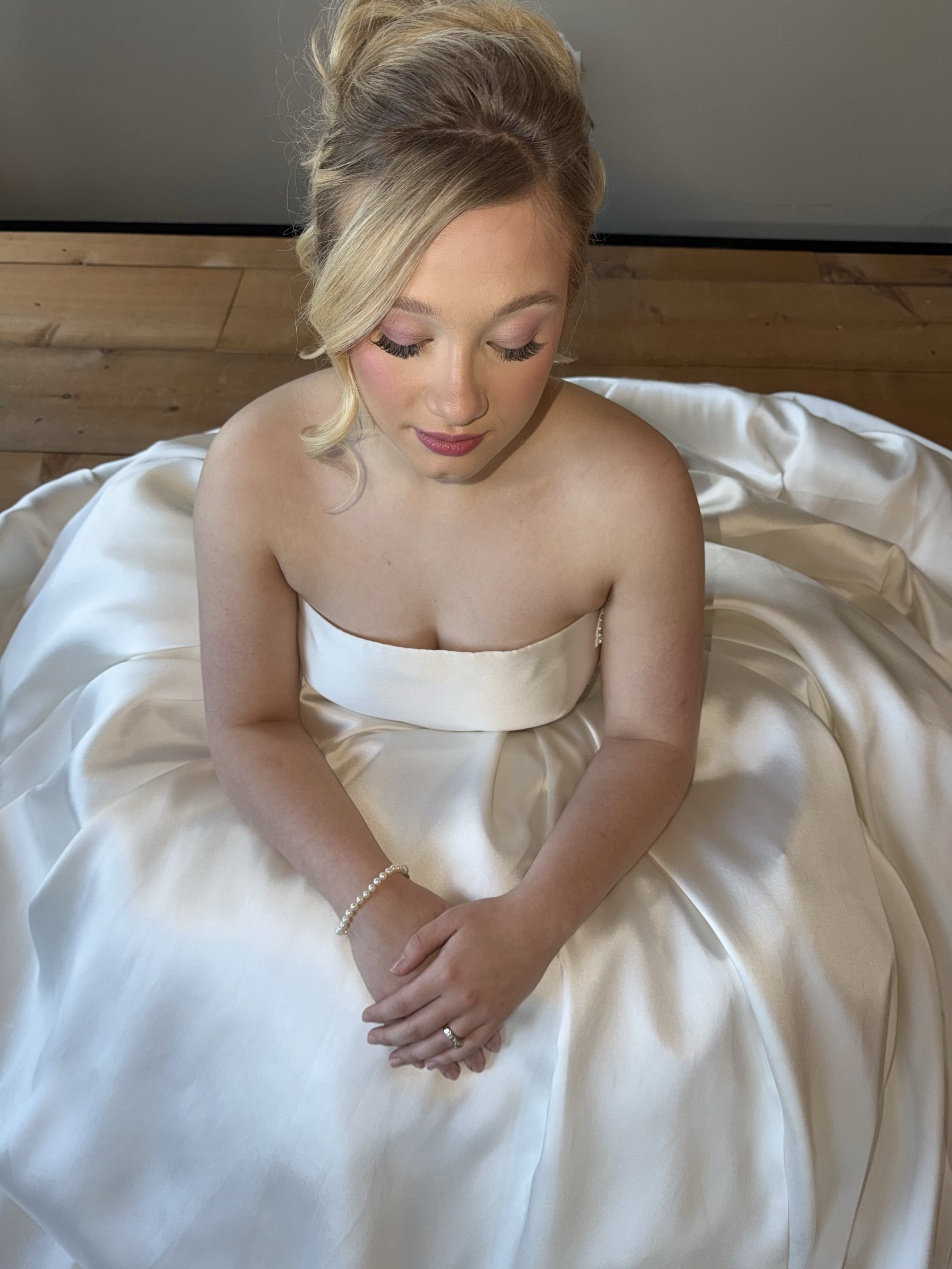 Brunette bride in white satin wedding gown sitting on the floor, with her hands clasped and eyes closed, wearing pearl bracelet and wedding ring.