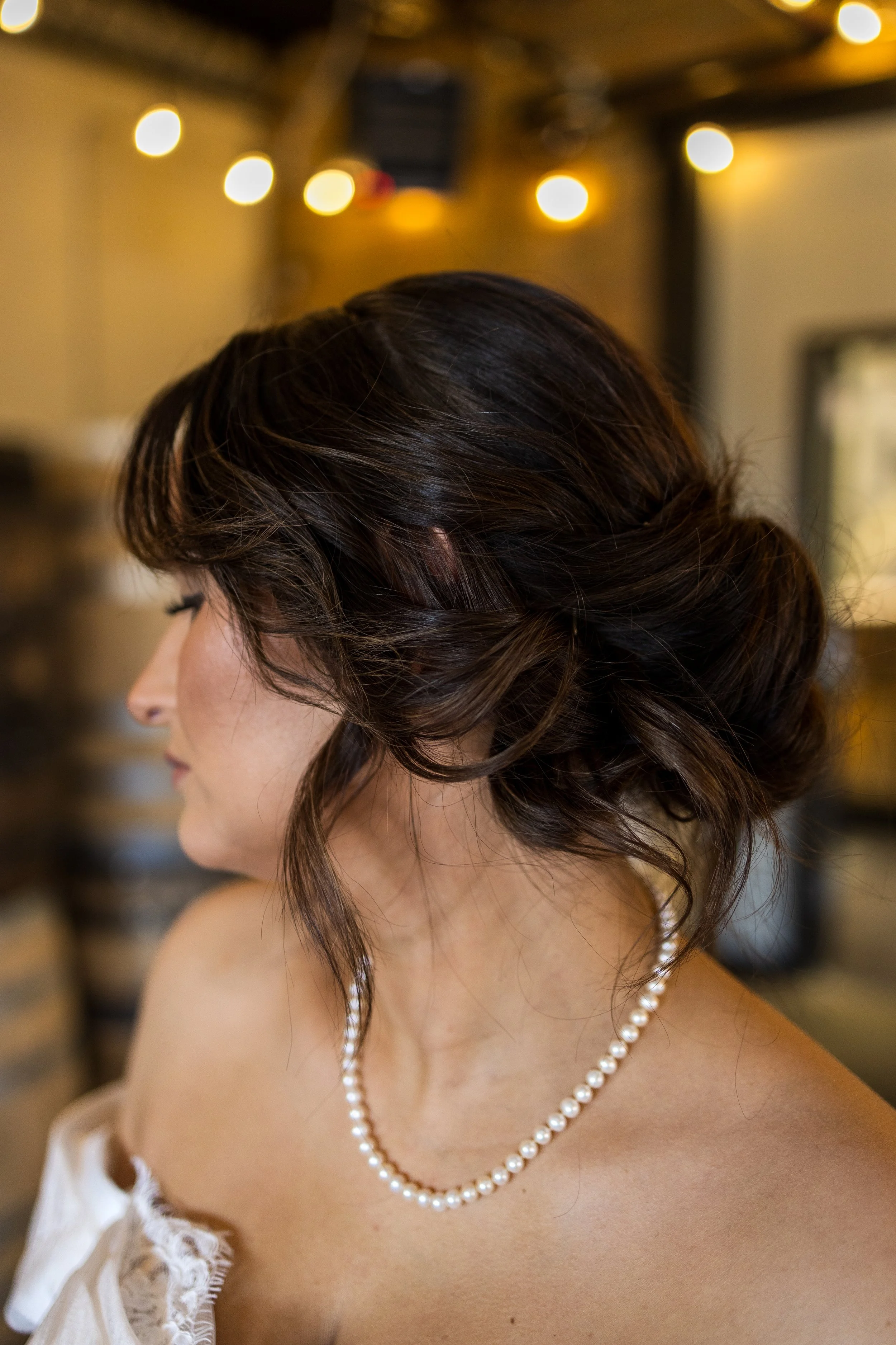 A woman with dark, wavy hair styled in an elegant updo, wearing a pearl necklace and a lace-off-shoulder dress, inside a warmly lit venue.