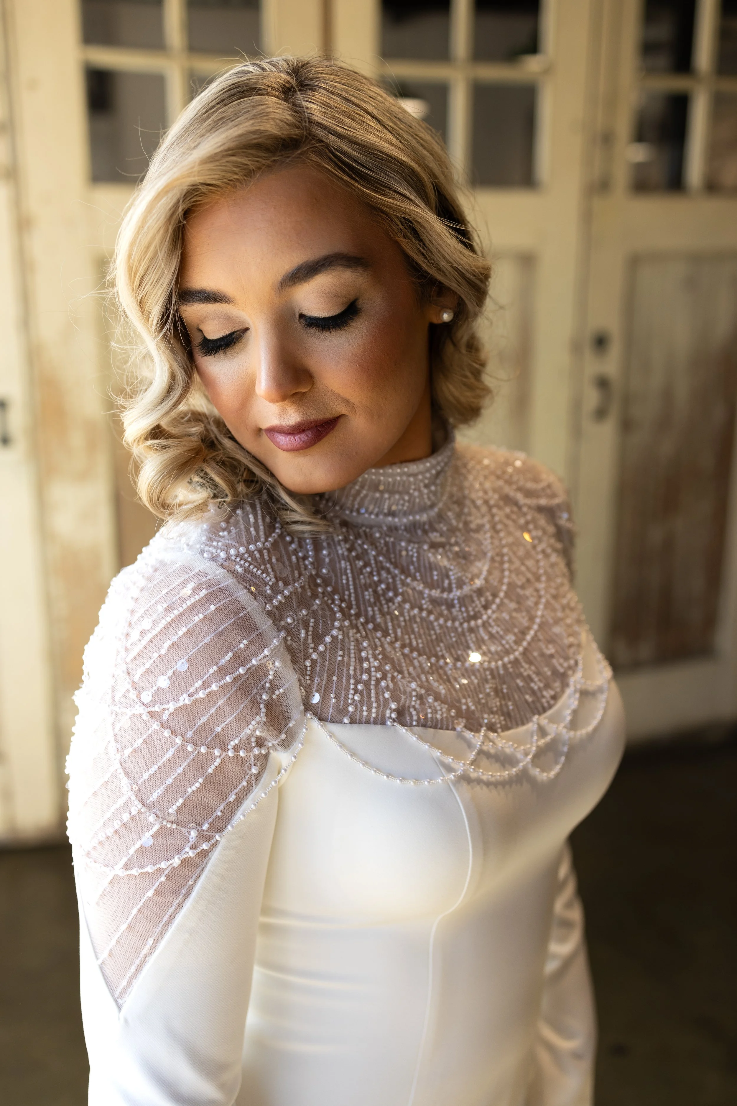 A woman with blonde, wavy hair and makeup, wearing a white dress with a high lace neckline featuring intricate beadwork and sequins, standing indoors in front of a rustic wooden door.