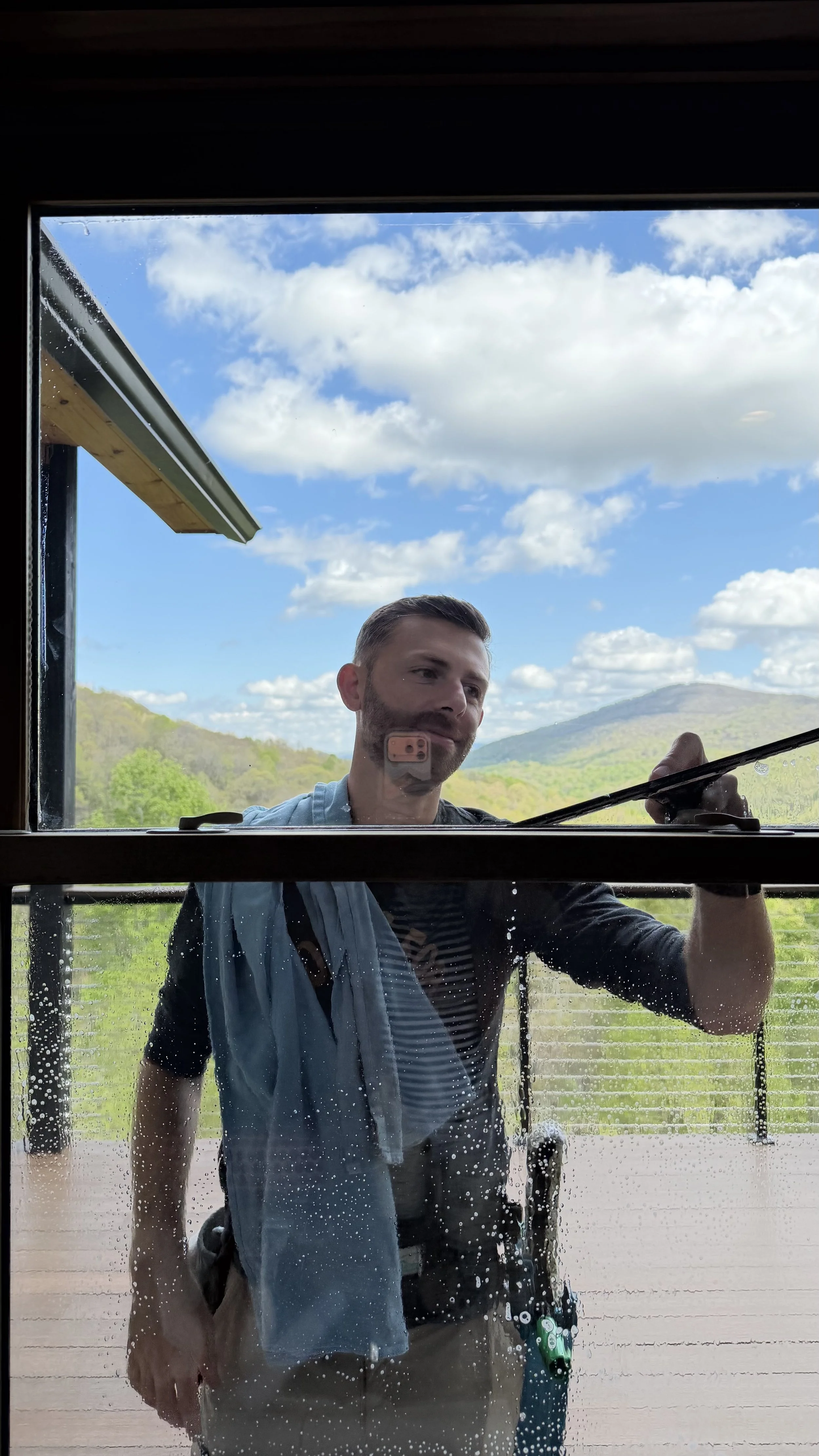 Man standing outside a building, seen through a window with water droplets, with a scenic landscape of hills and blue skies with clouds in the background, holding a cleaning tool. cleaning windows in Boone, NC