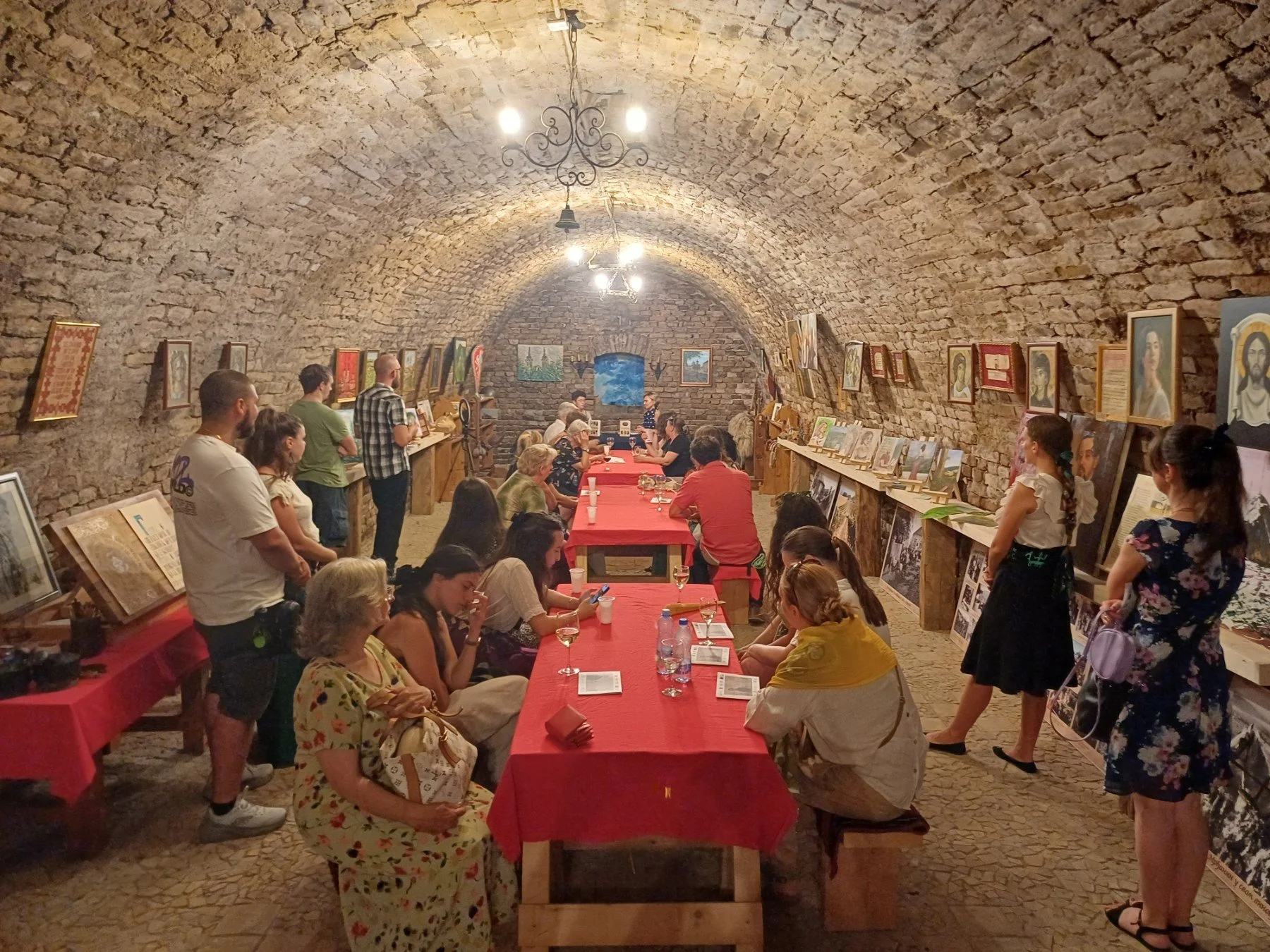 A group of people gathered in a brick underground art gallery with paintings on the walls. They are seated at red-covered tables and some are standing, listening to a speaker near the paintings. The ceiling is vaulted with exposed brick, and there are chandeliers lighting the room.