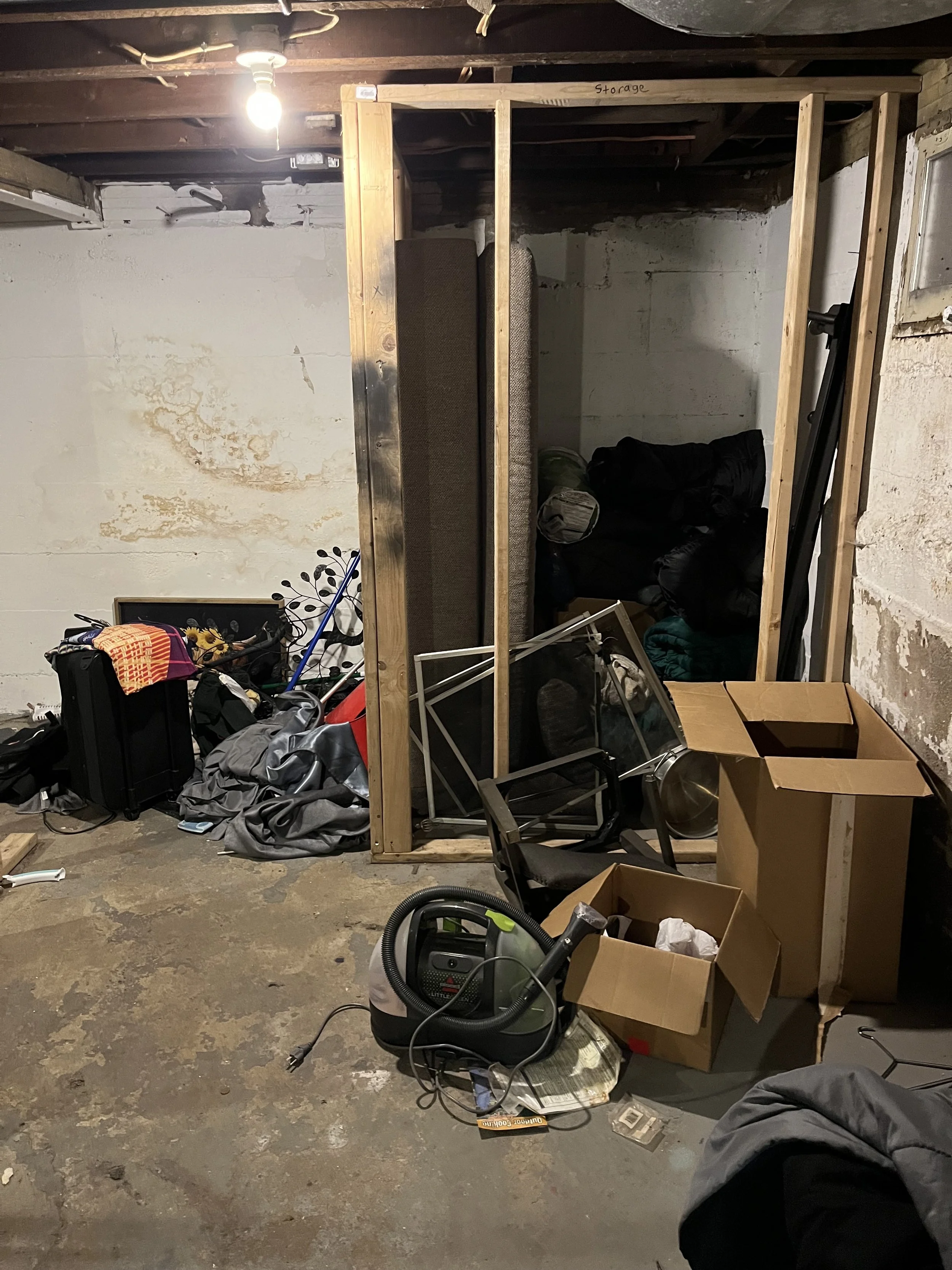 Unfinished basement with various clutter, including boxes, a vacuum cleaner, chairs, a framed picture, and stored items, with exposed wooden framing and a single light bulb overhead.