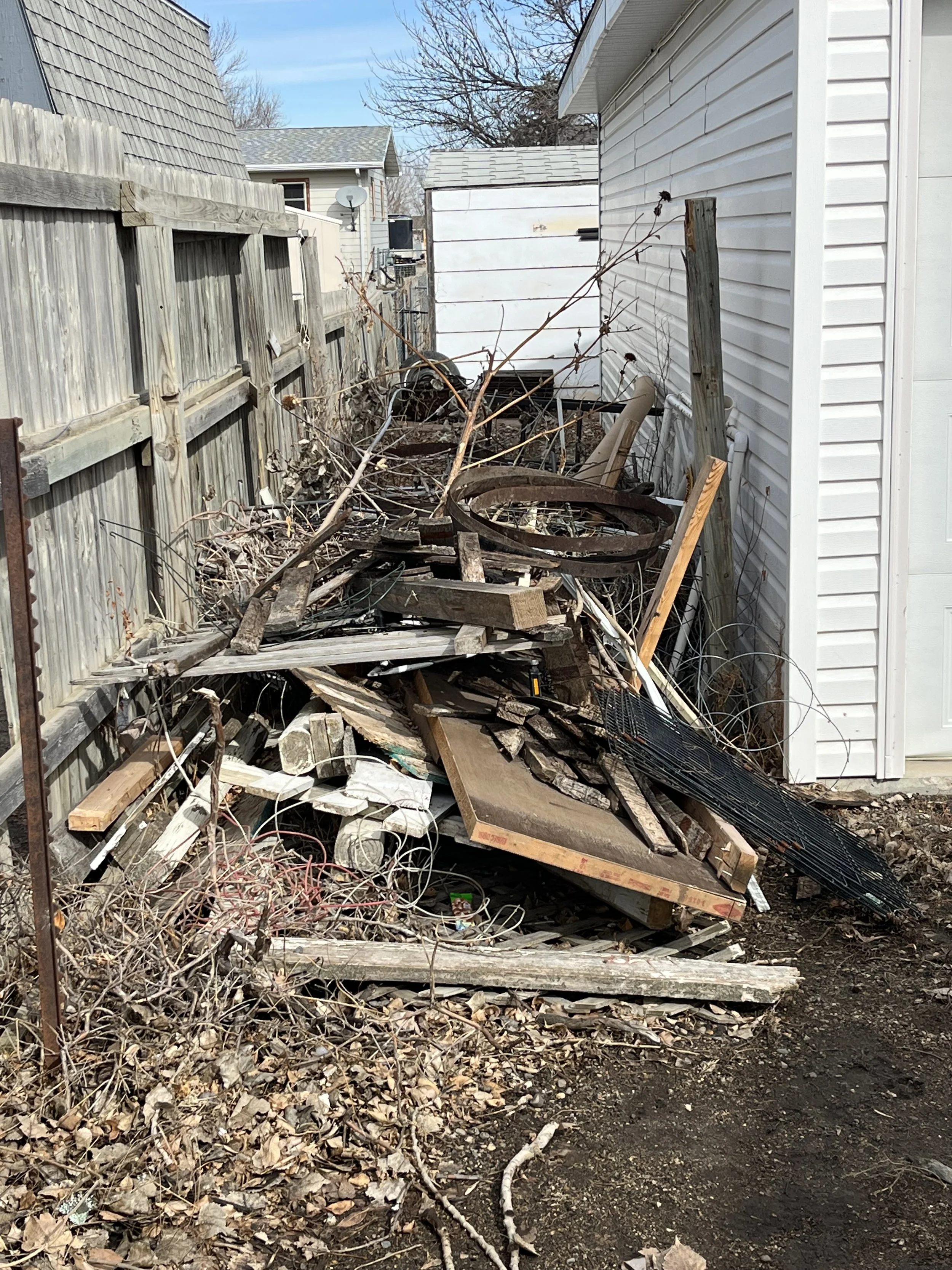 Piles of scrap wood, metal, and debris stacked along a narrow outdoor space between a wooden fence and a white house, with bare branches and scattered leaves on the ground.
