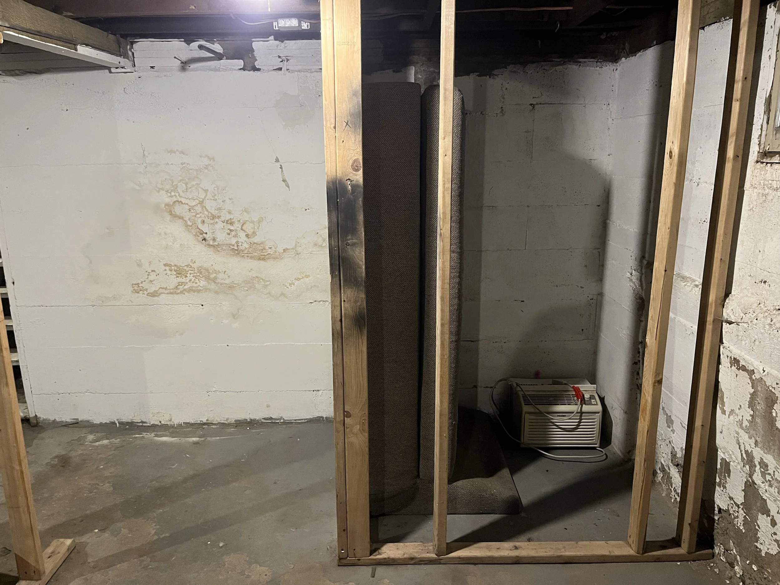 A partially constructed small room or closet with wooden framing, a carpeted box or piece of furniture inside, and a portable heater on the floor in a basement with unfinished walls and concrete floor.