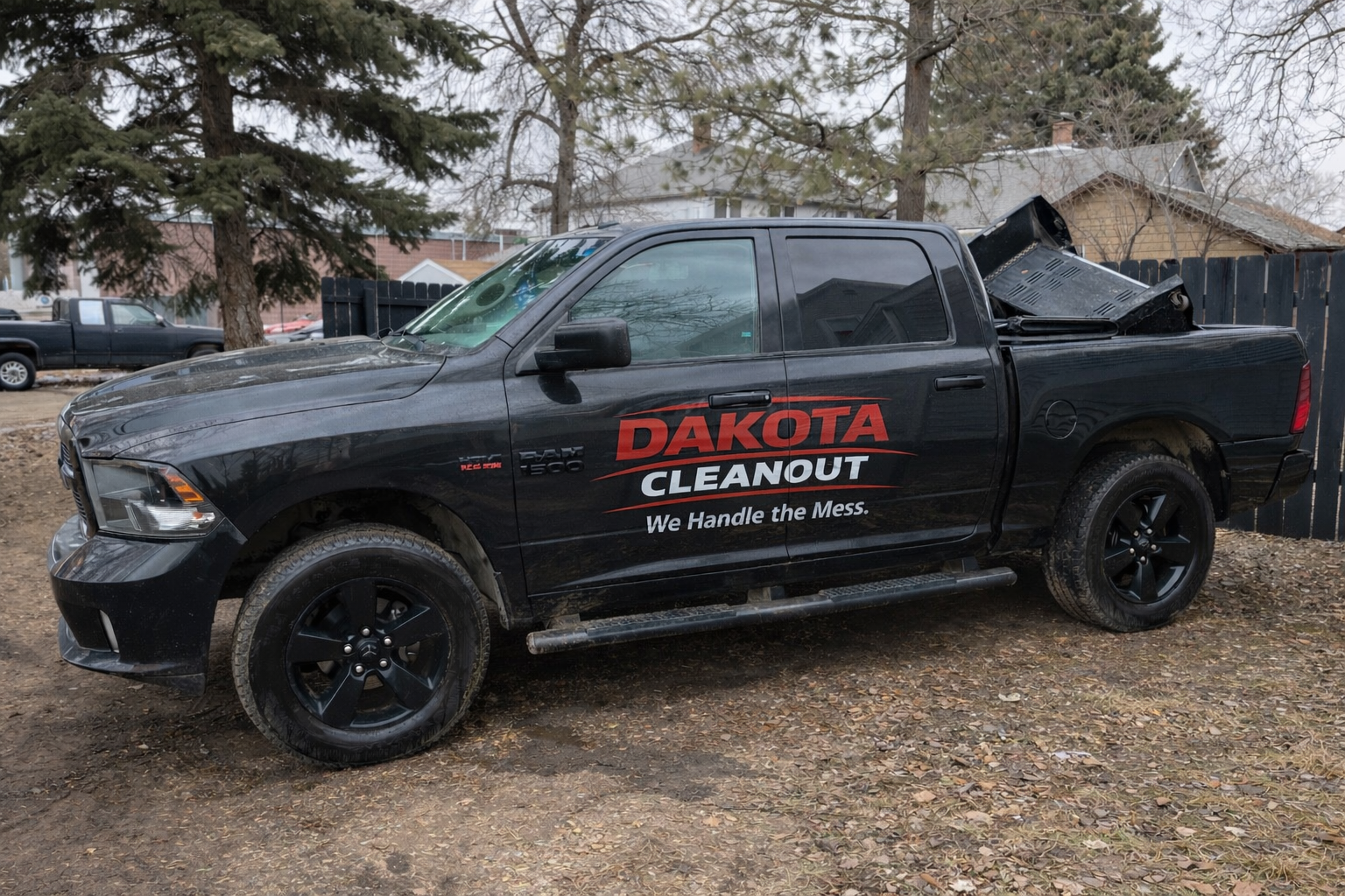 Black pickup truck with 'Dakota Cleanout' logo and slogan parked on dirt lot with fallen leaves, trees, and houses in background.