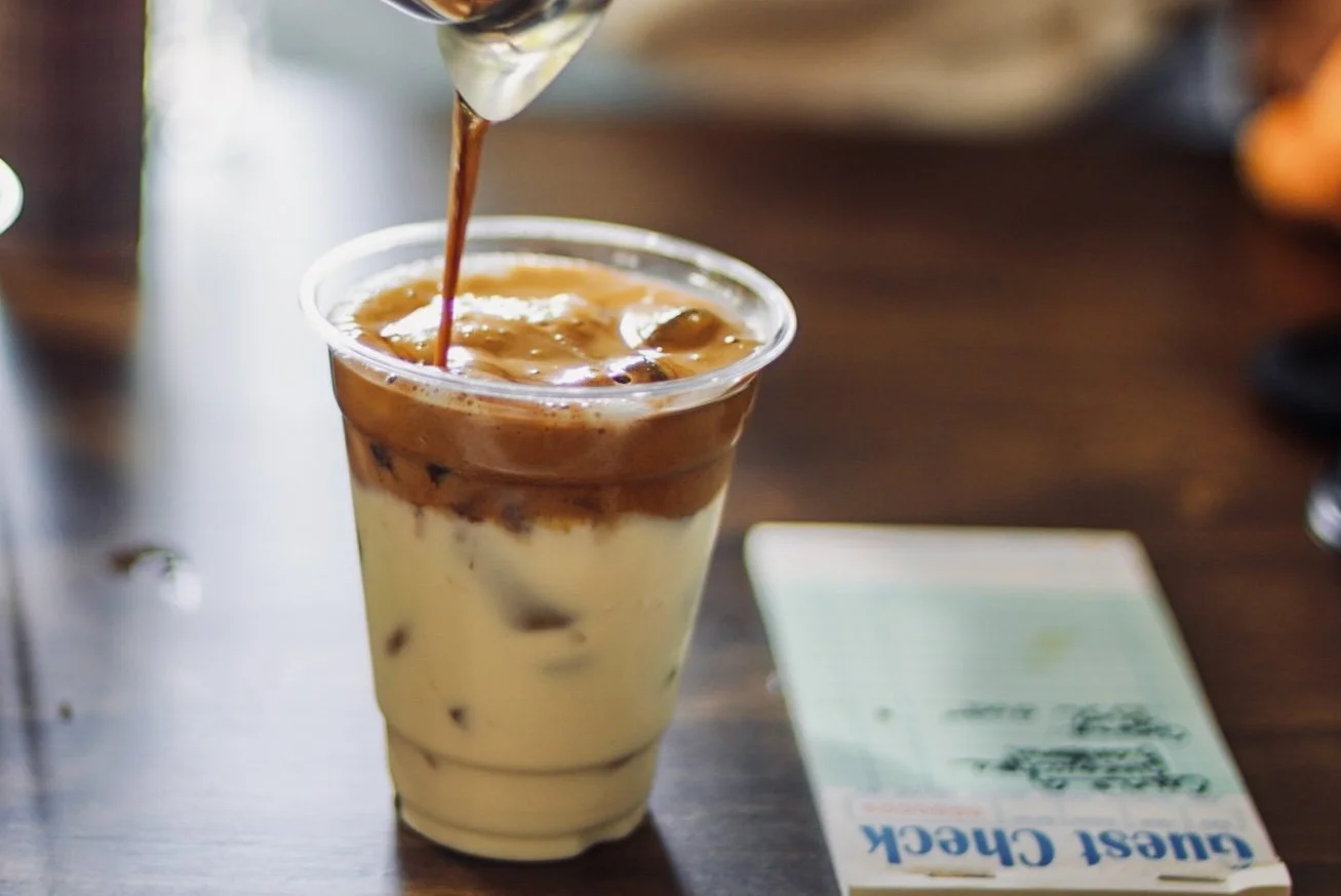 A glass of iced caramel macchiato being poured with espresso on top.