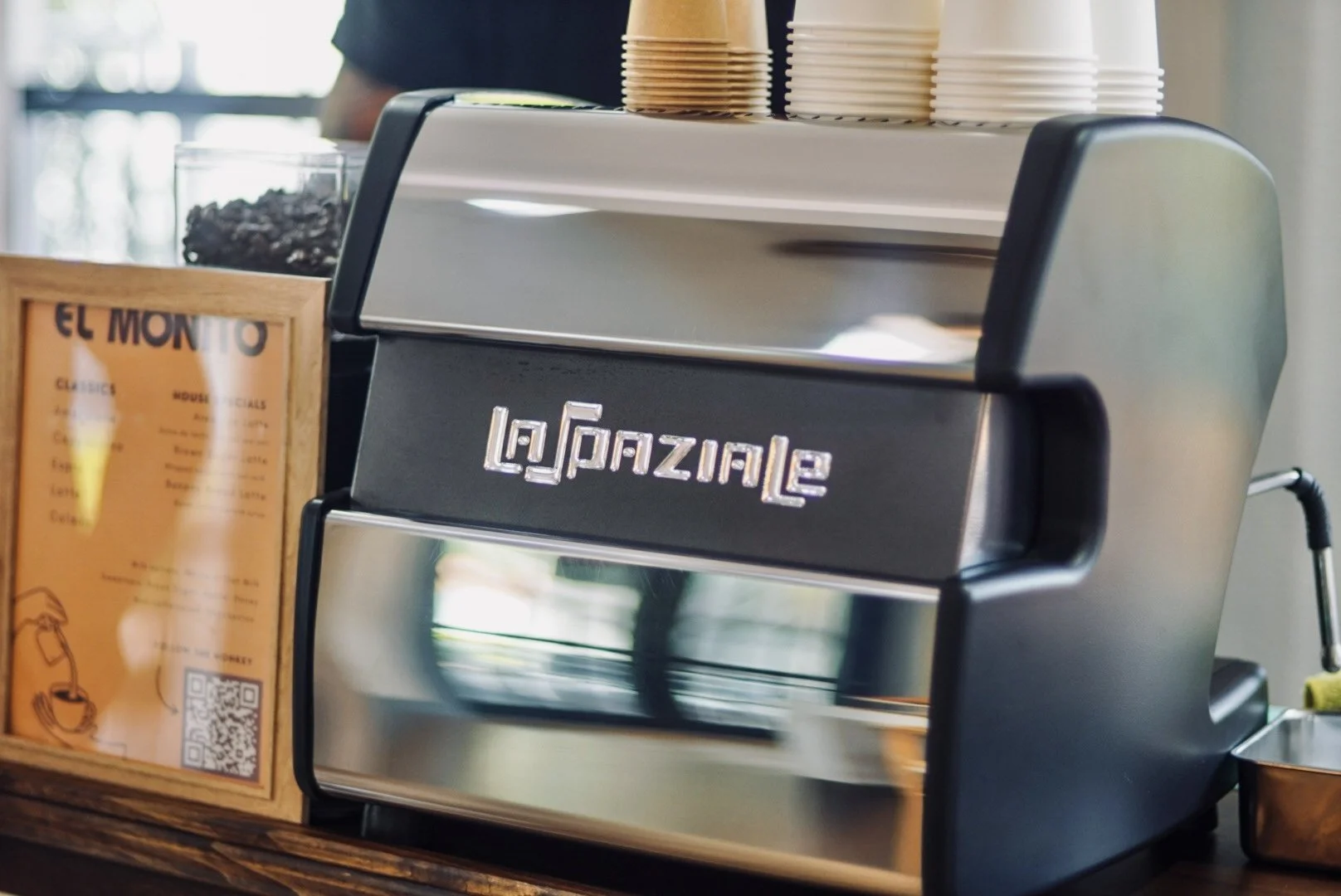A sleek, modern espresso machine with the brand name 'La Marzocco' on the front, placed on a wooden bar or countertop. In the background, stacked cups and a sign with menu options are visible.