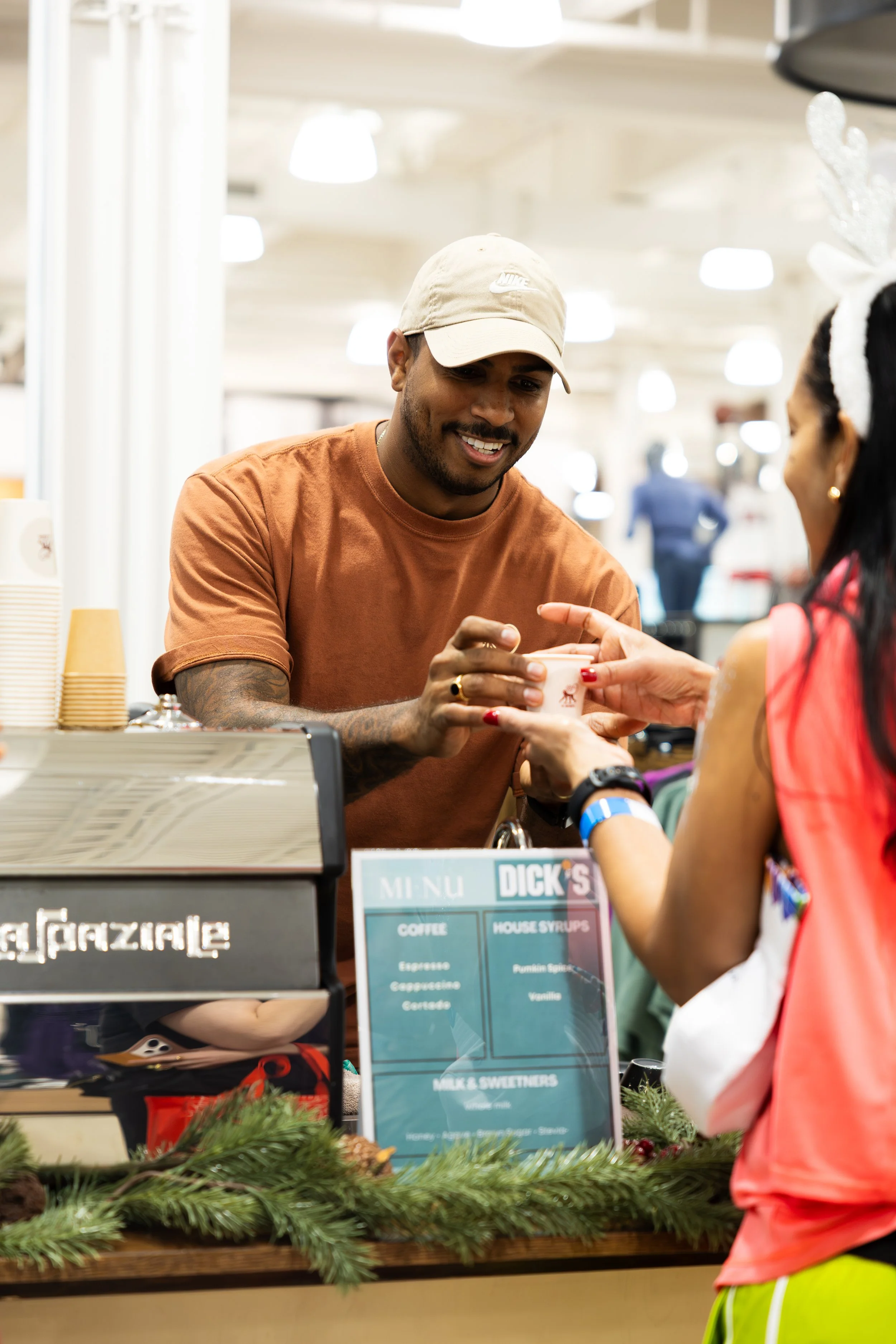 A man at a coffee stand is handing a cup to a woman. The scene suggests a holiday or festive setting, with holiday greenery on the counter and a woman wearing reindeer antlers.