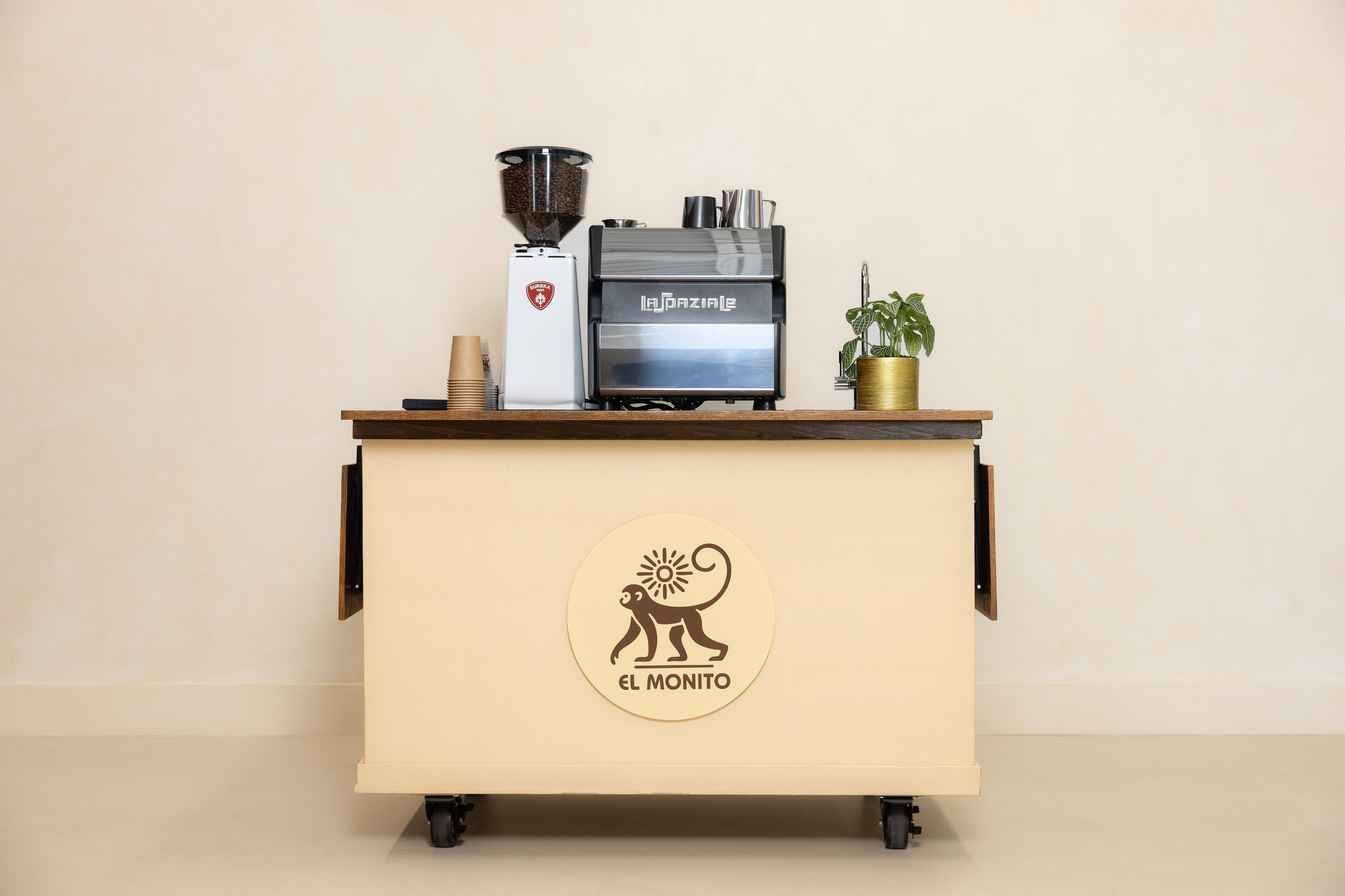 Coffee station with espresso machine, coffee grinder, disposable cups, and a potted plant, behind a cart with a logo of a monkey and the words 'El Monito'.
