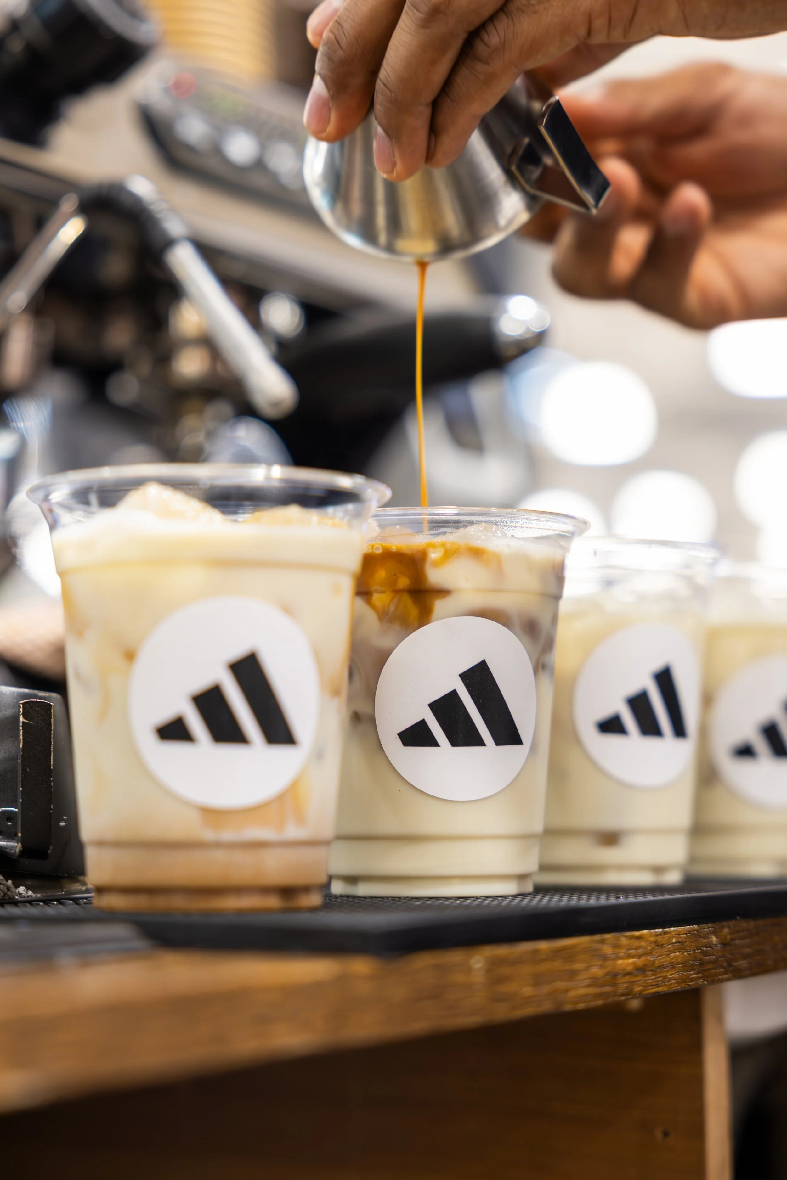 A barista pours espresso into a cup of iced coffee. Several cups of iced coffee with a logo are lined up on a wooden counter.