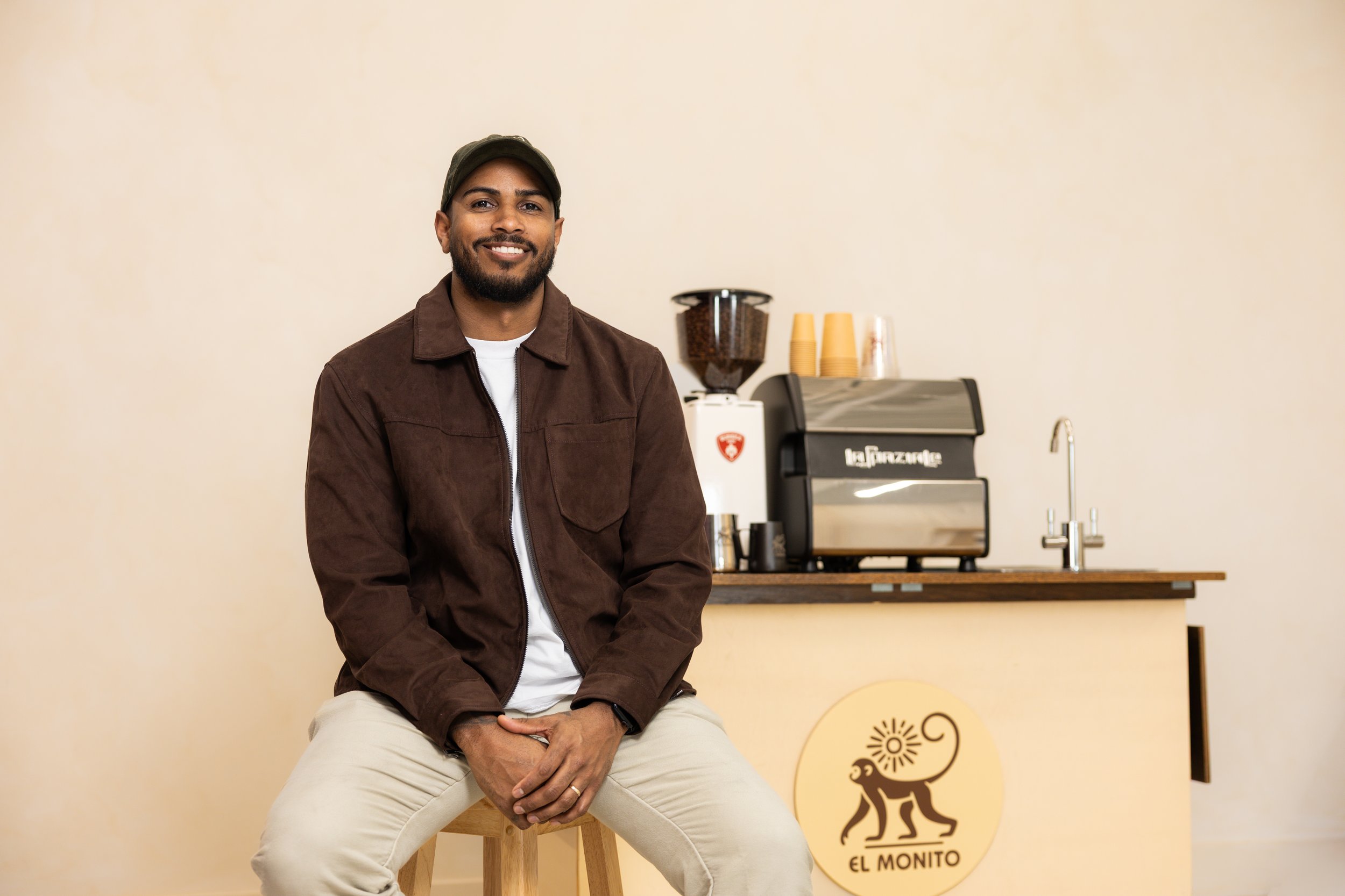 Man sitting on a stool in front of a coffee station with a coffee grinder and espresso machine, in a coffee shop called El Monito.